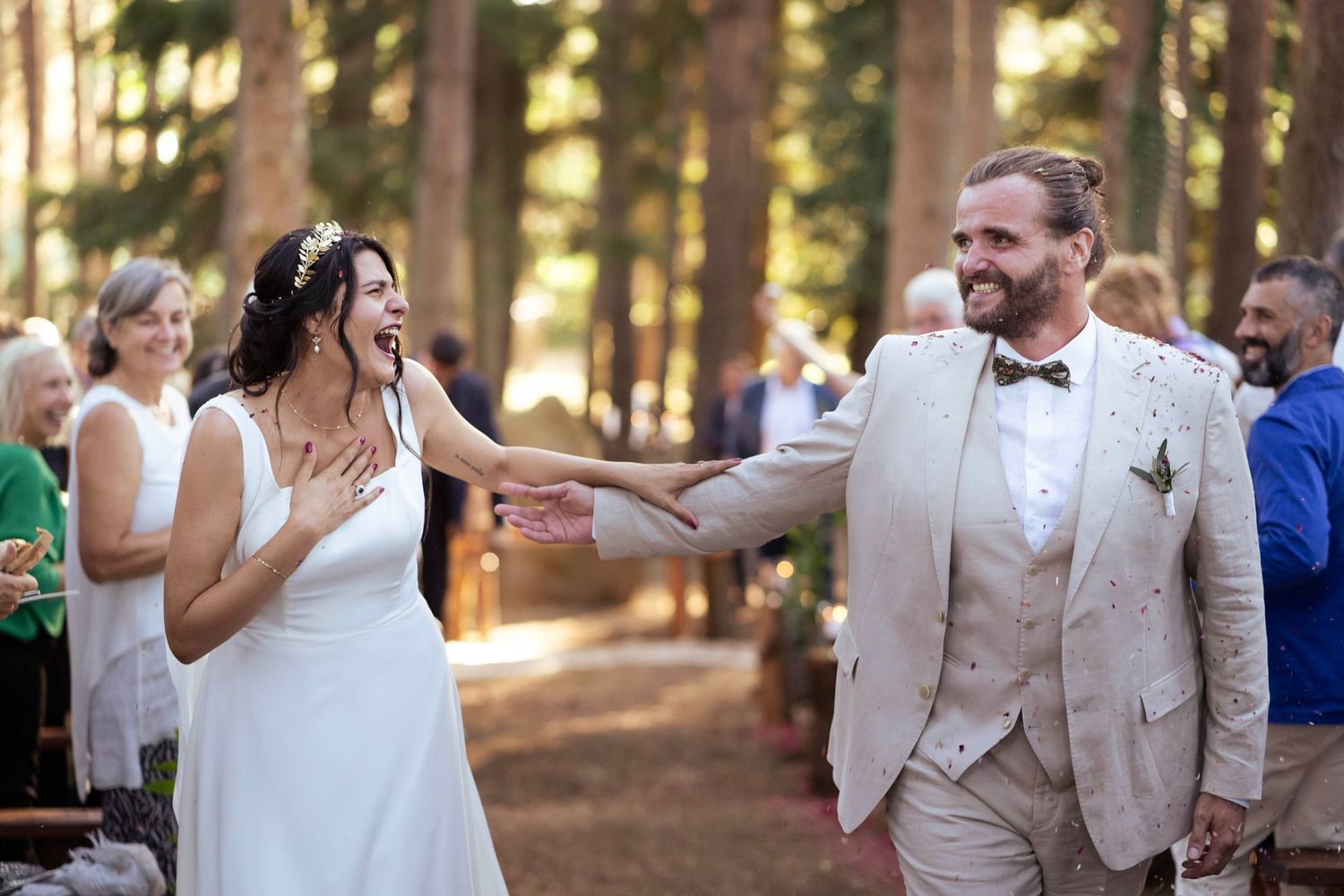A joyful wedding celebration in a sunlit forest with guests looking on as newlyweds share a playful moment.