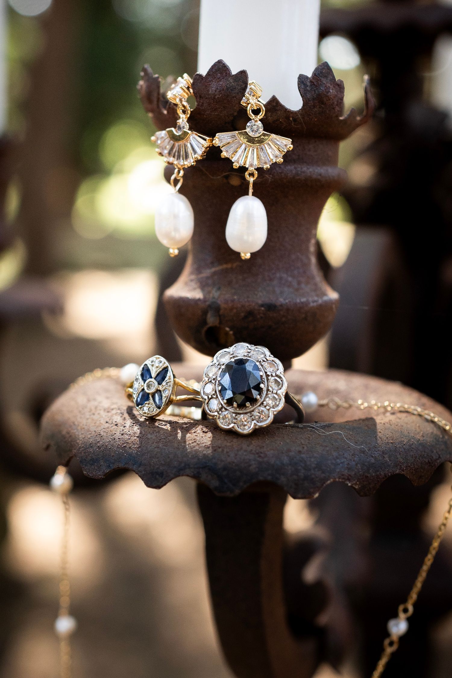 Pearl drop earrings and sapphire rings displayed on antique dark metal candlestick.