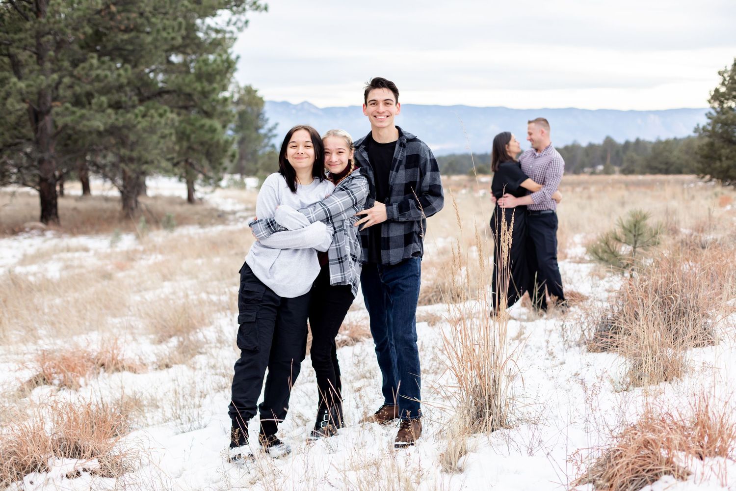 Three teen children hug and smile while their parents embrace in the background in Black Forest, Colorado.