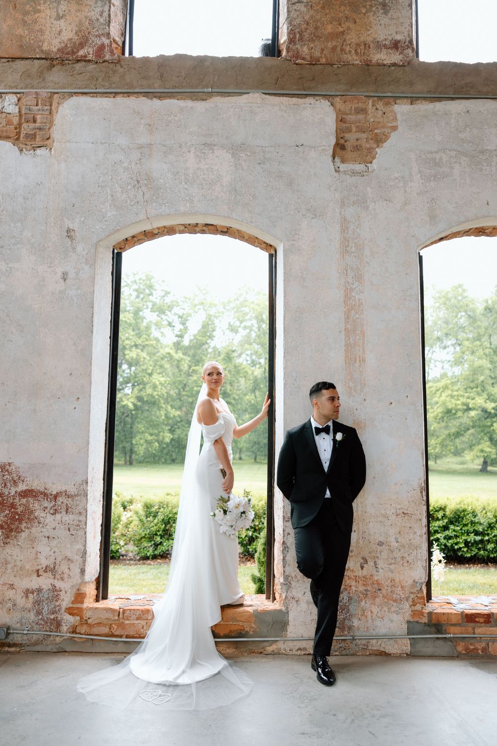 Bride and groom stand separated in rustic archways with greenery backdrop.