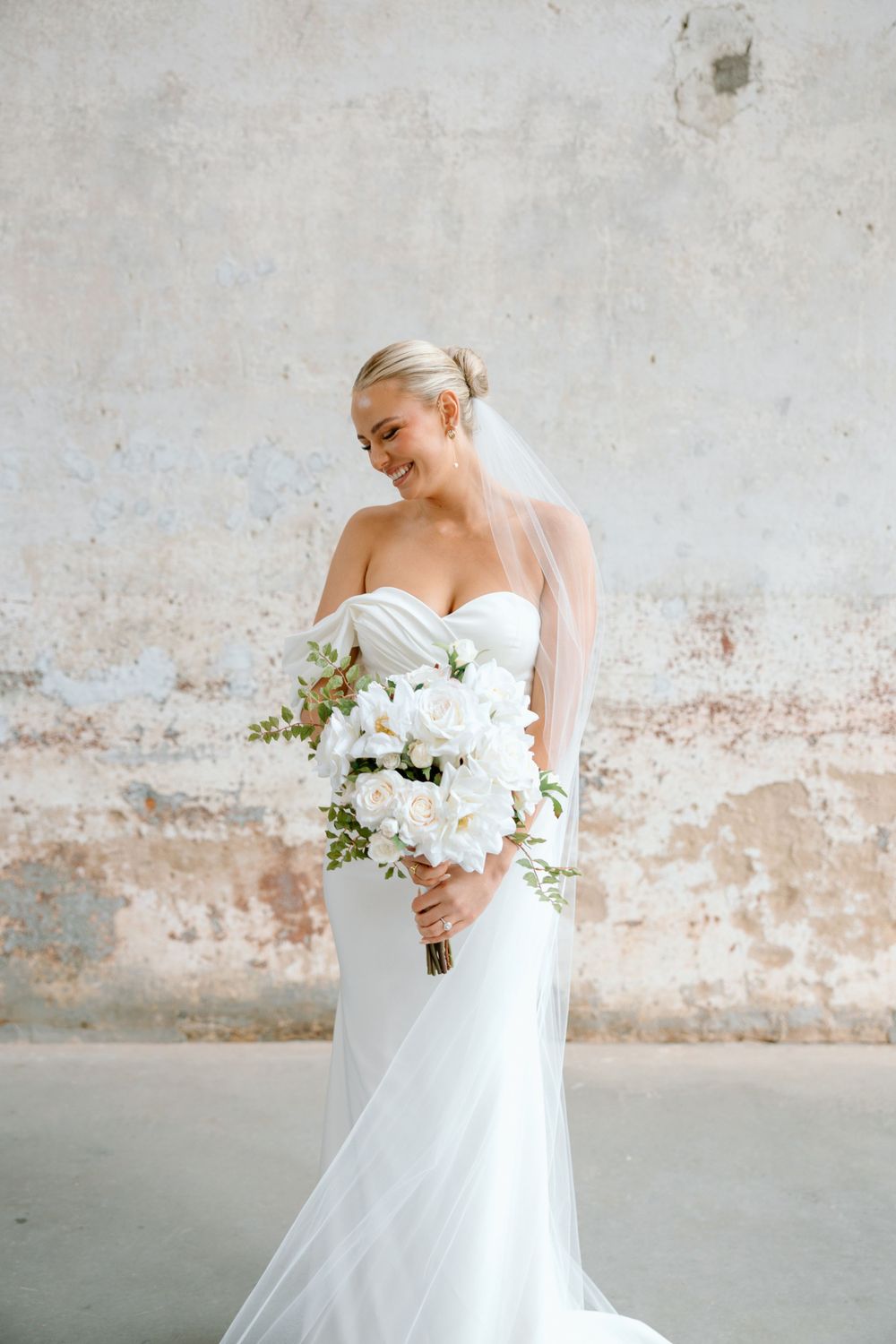 Bride in strapless gown holds cascading white bouquet against rustic wall.