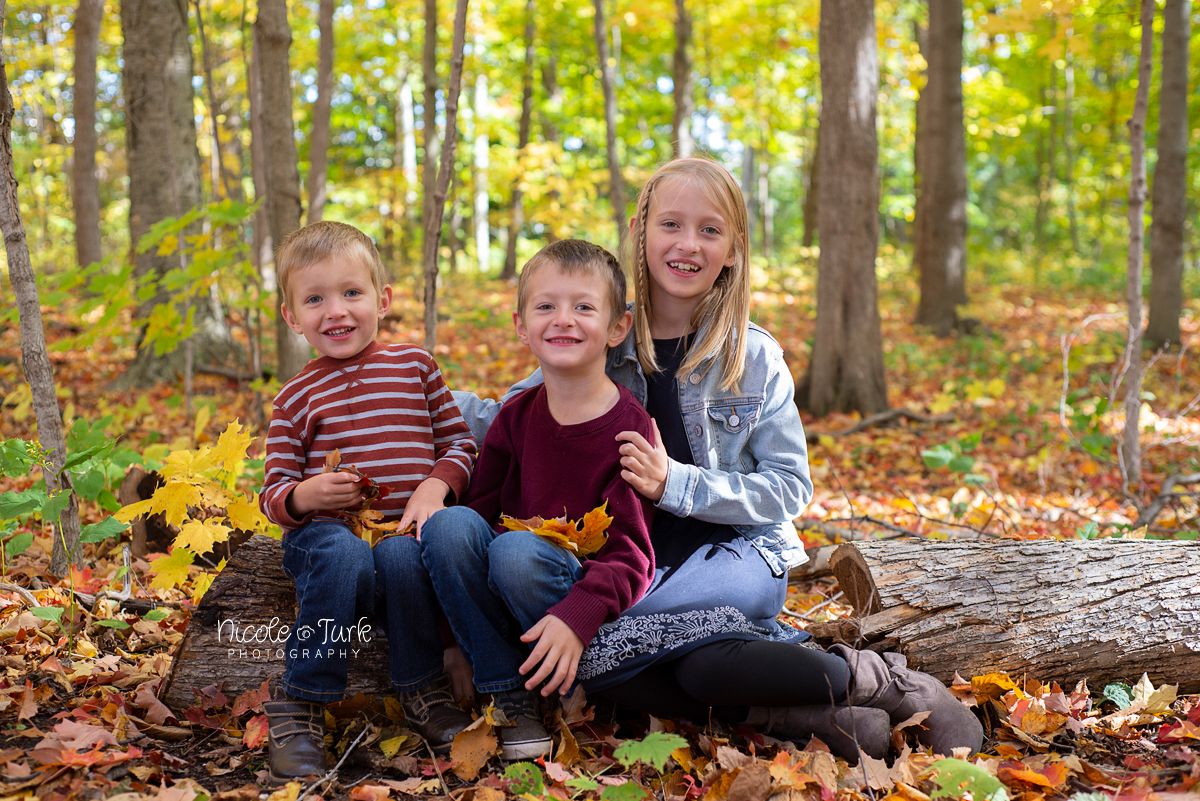 3 young happy kids pose together for a photo in the fall leaves in Courtice while sitting on a log