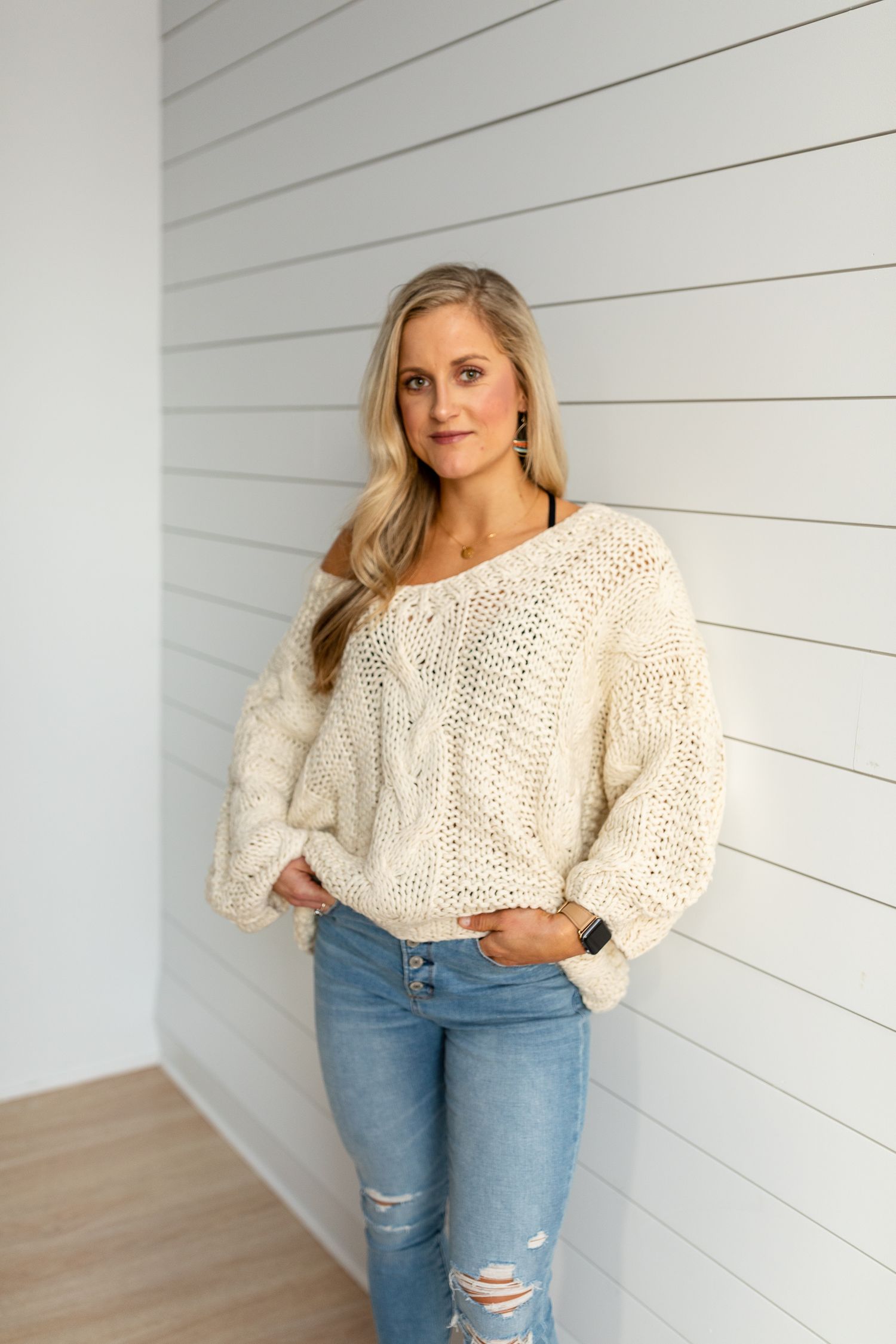 Woman in cream cable knit sweater and distressed jeans against white shiplap wall.
