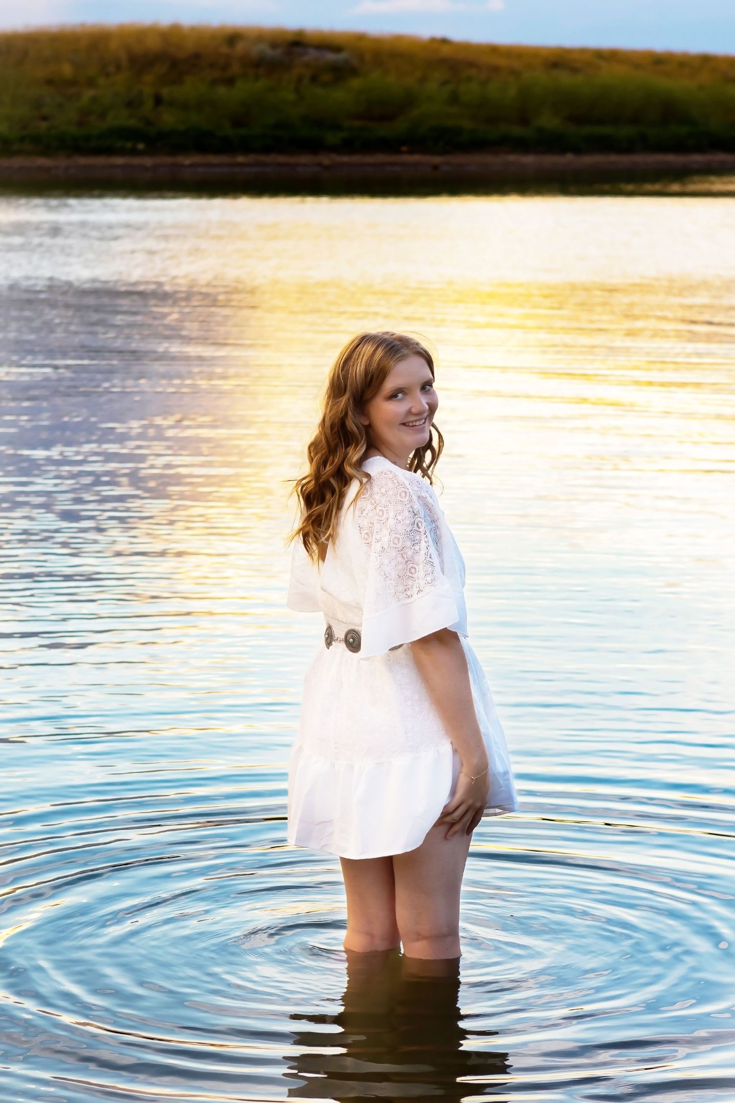 A person in a white dress stands in shallow water during golden hour at a lake.