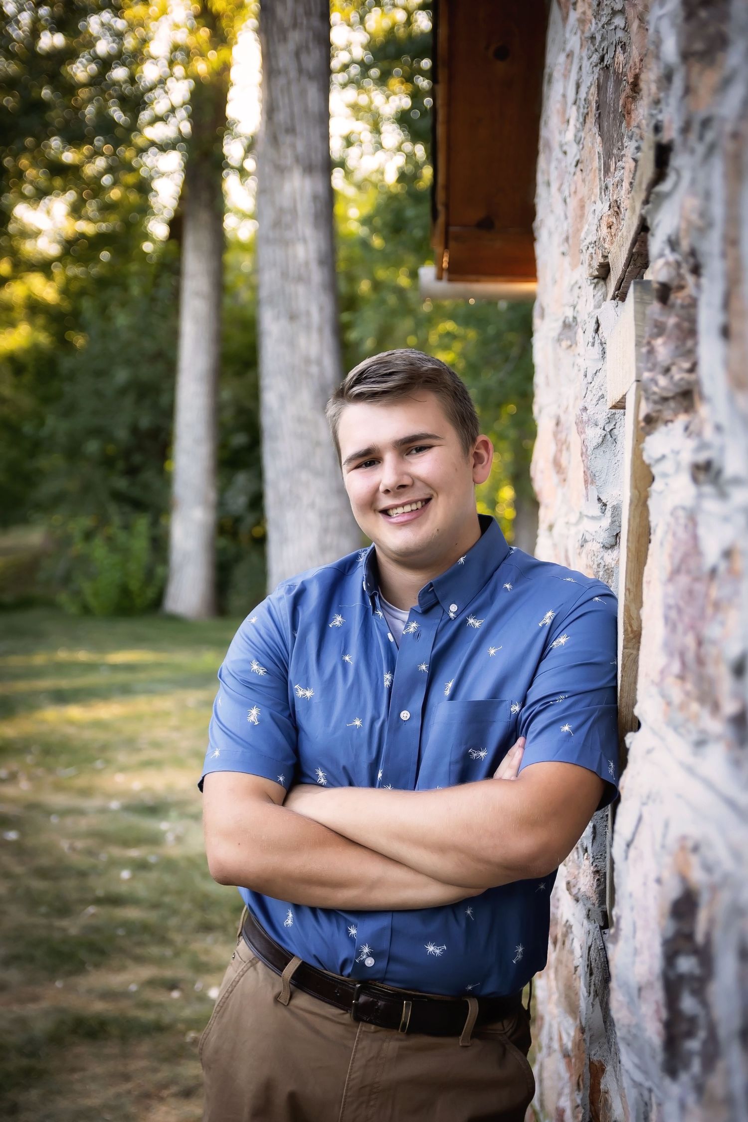 Professional portrait of someone in a blue button-down shirt leaning against a rustic stone wall at golden hour.