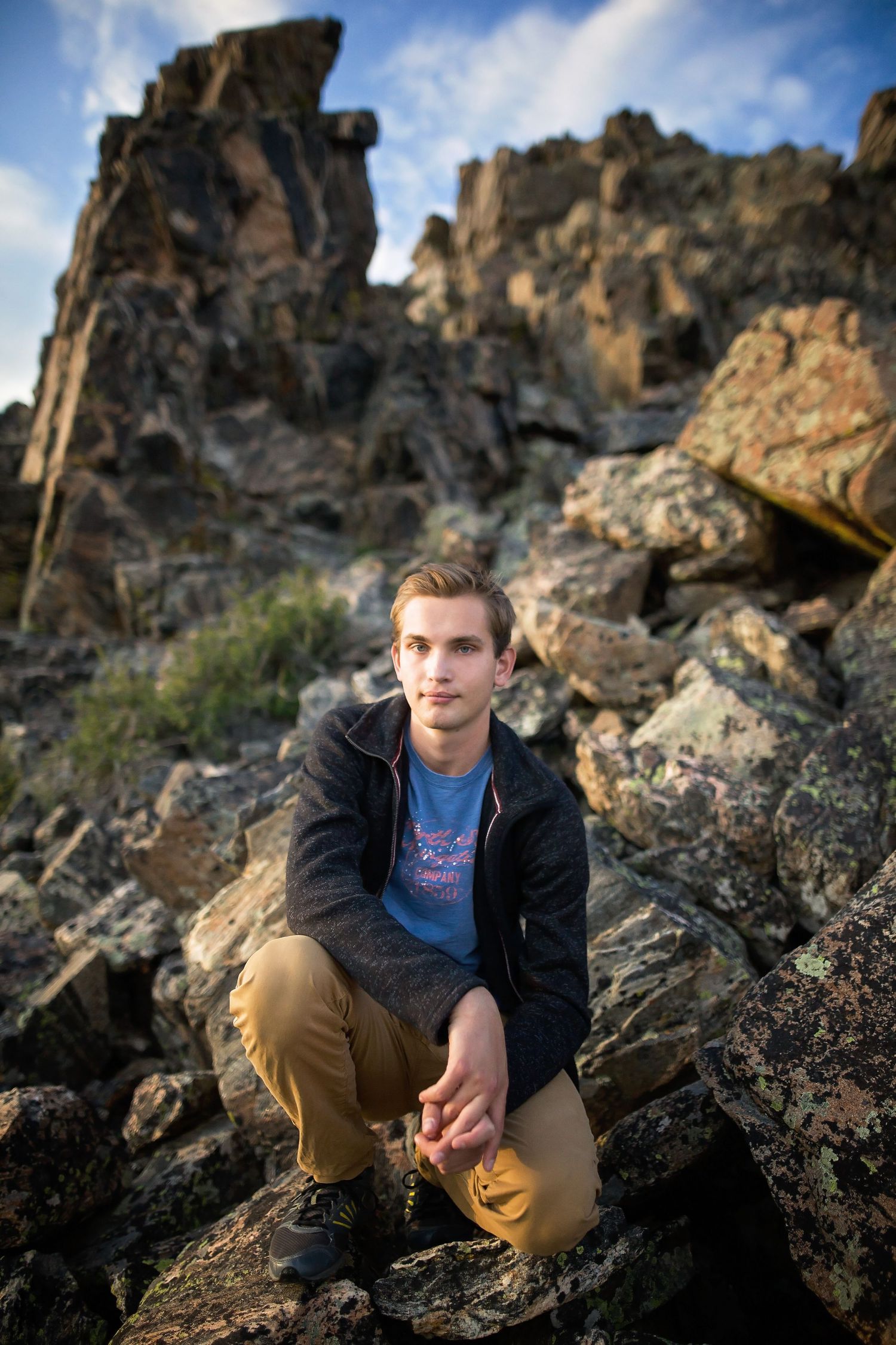 Person crouching on rocky outcrop with dramatic cliffs and blue sky in background.