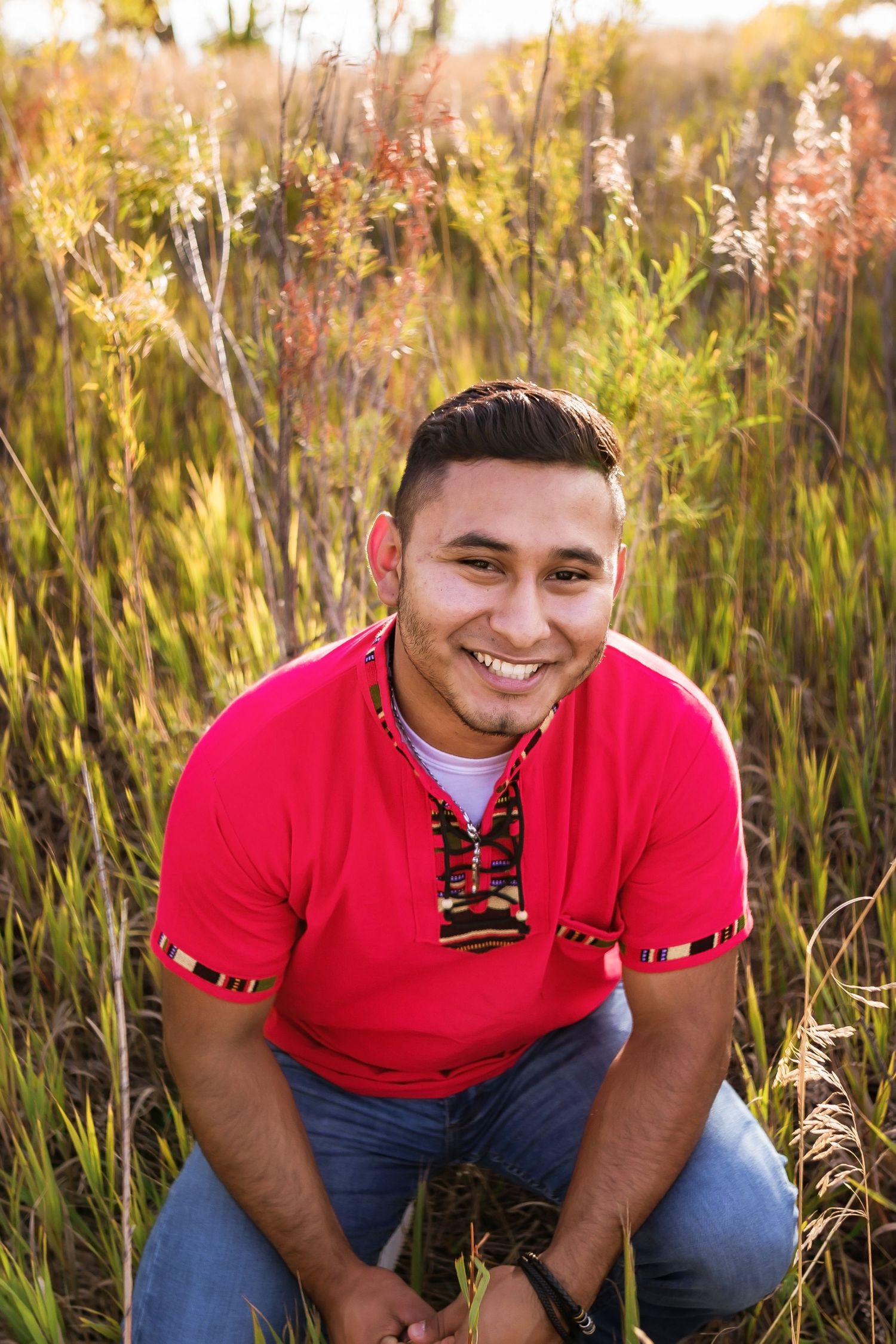 A person in a red shirt poses outdoors among tall grass during golden hour.