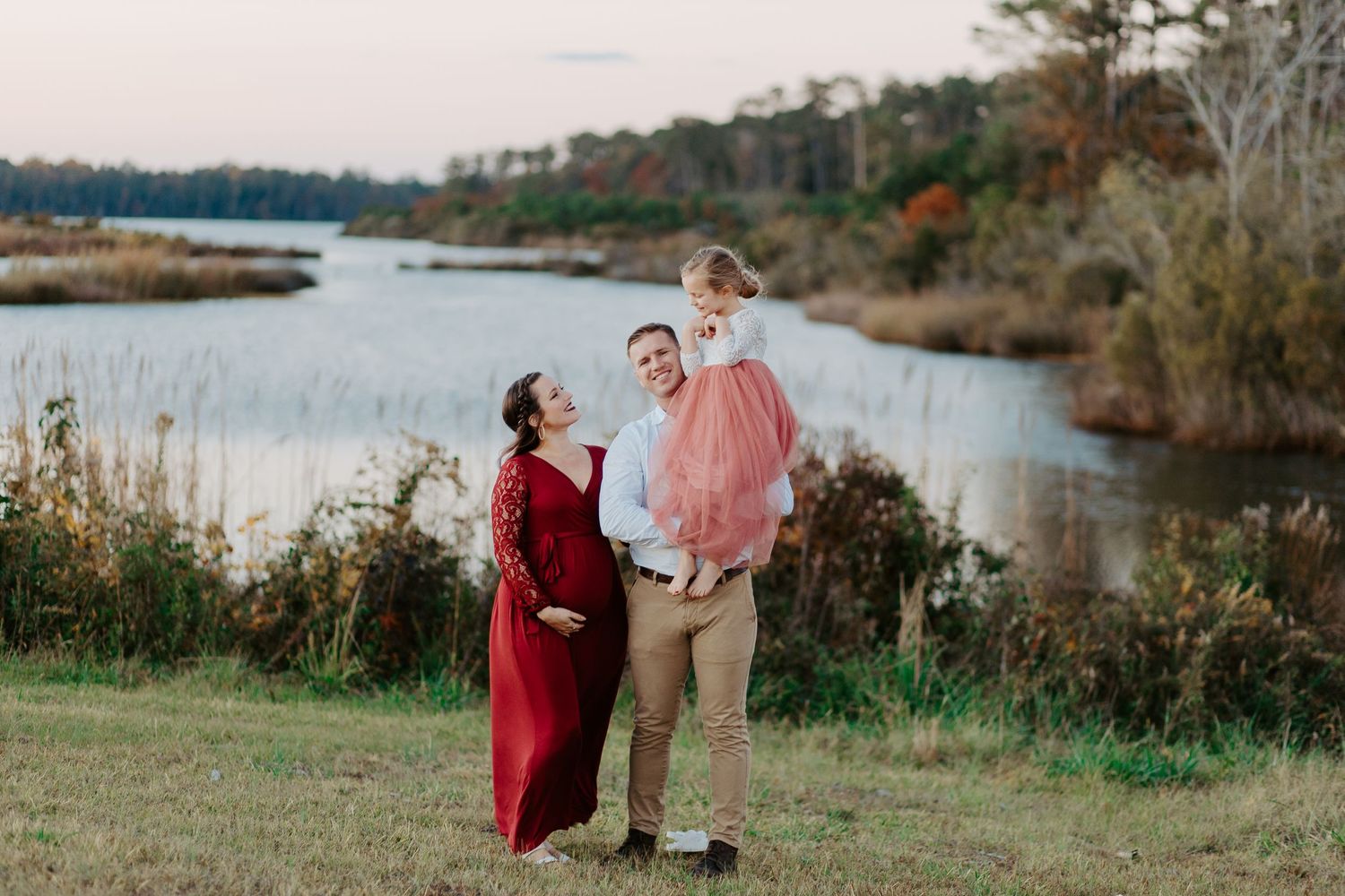 Pregnant woman in red gown with husband holding toddler near a scenic lake in autumn landscape.