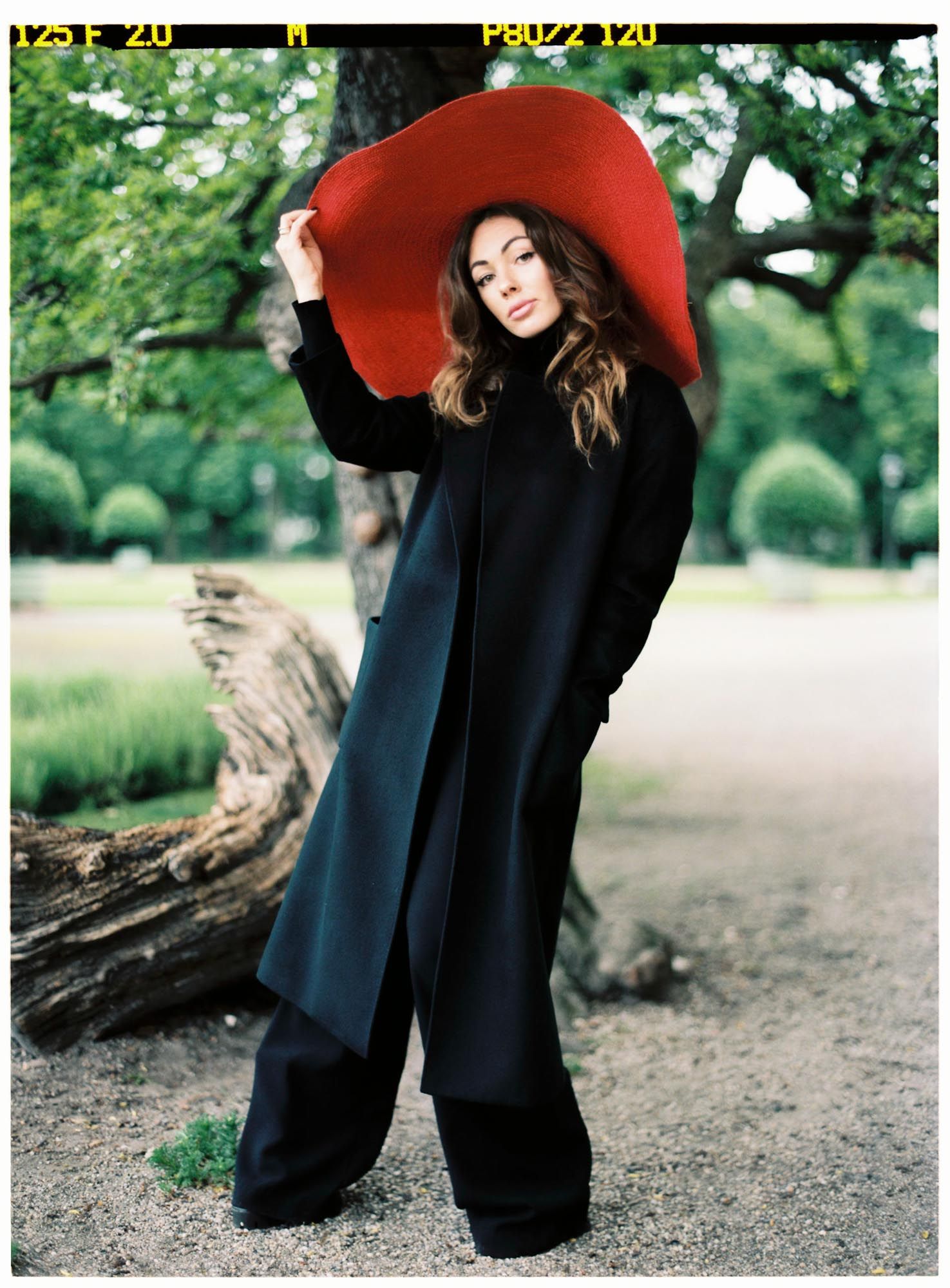 Fashionable figure in black coat and dramatic red wide-brimmed hat poses in outdoor garden setting.