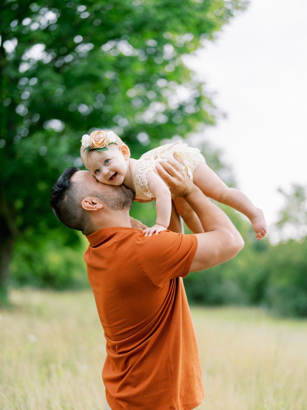 Parent playing with toddler in white dress outdoors on sunny day.