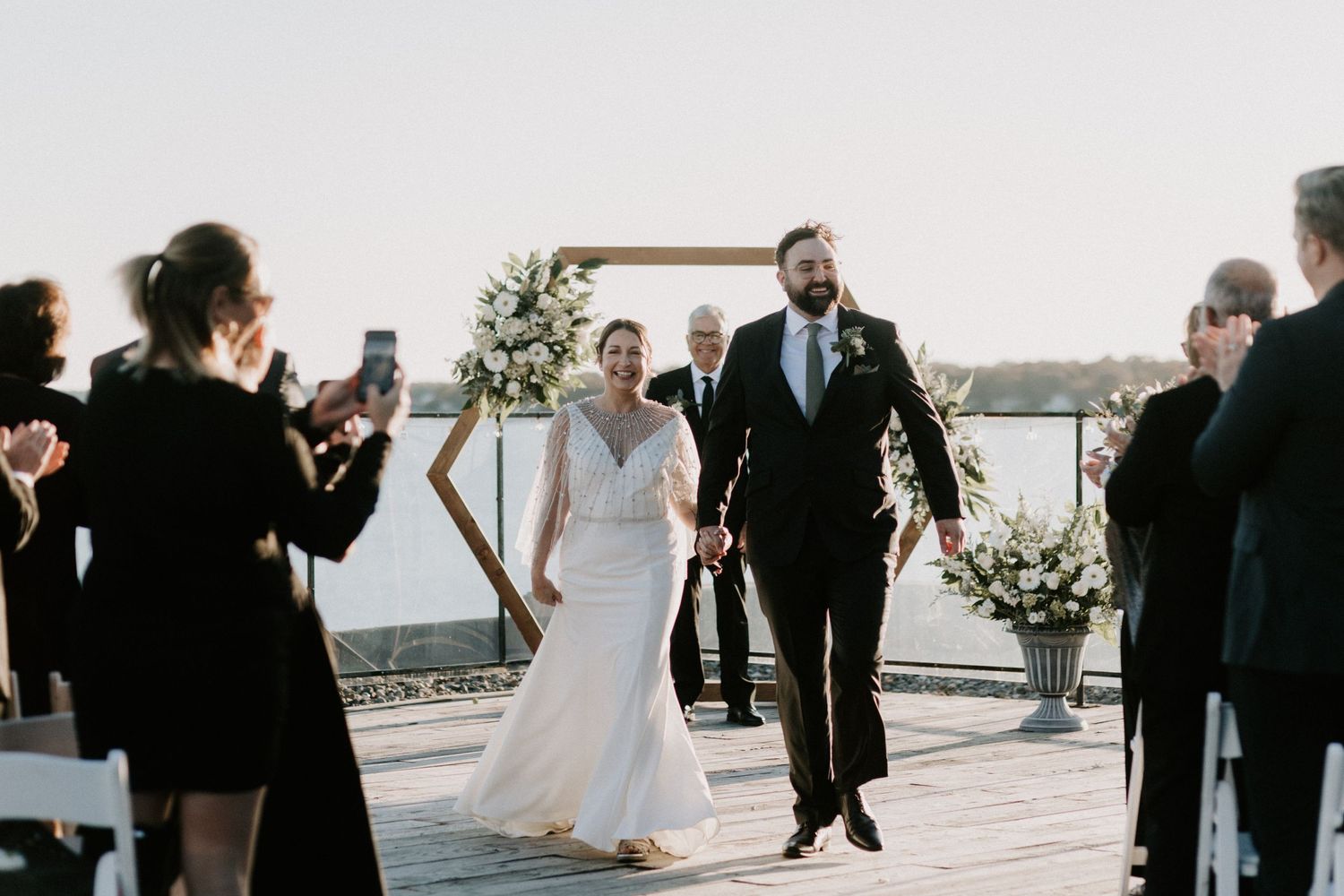 Newlyweds walk down the aisle on a waterfront deck ceremony with floral arch and cheering guests at sunset.
