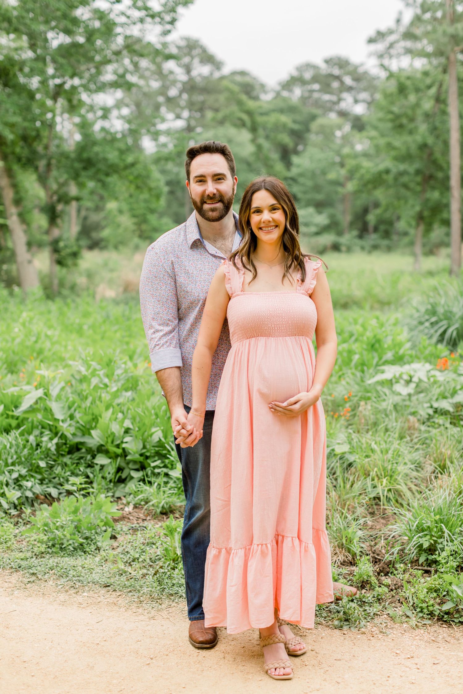 A couple holds hands while posing on a dirt path surrounded by green foliage.