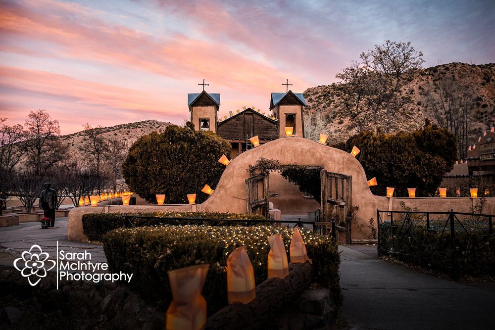 Historic Santuario church at sunset with pink sky and luminarias along pathway.