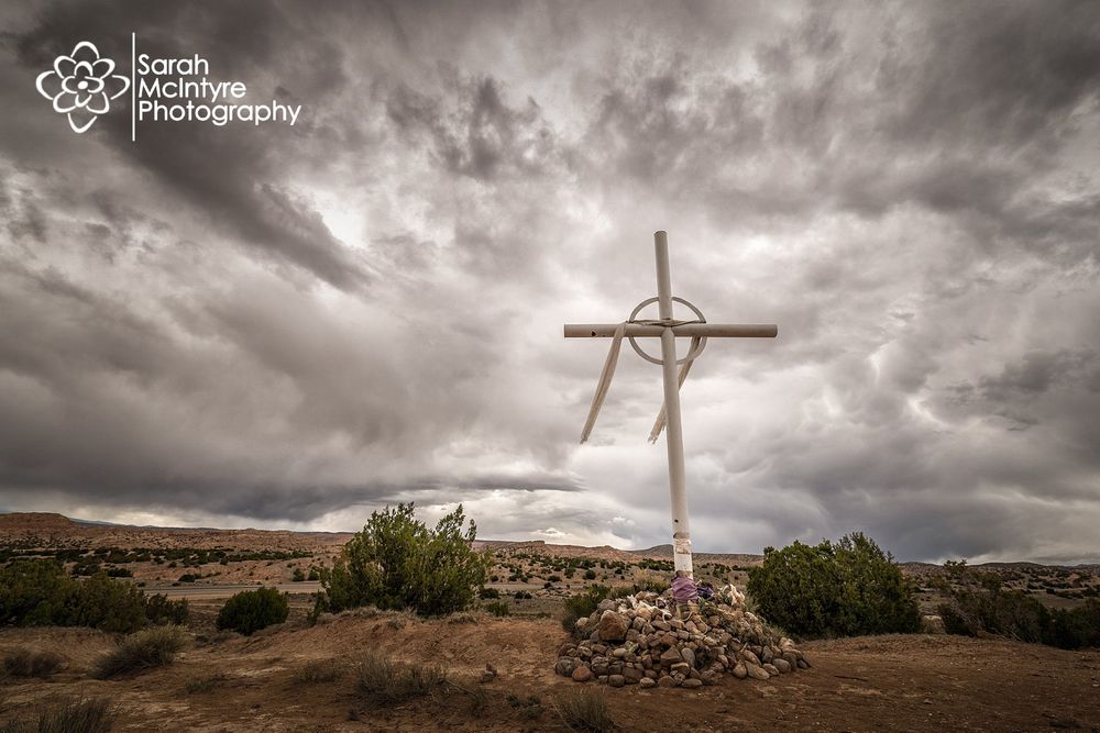 Wooden cross silhouetted against dramatic stormy sky in desert landscape.
