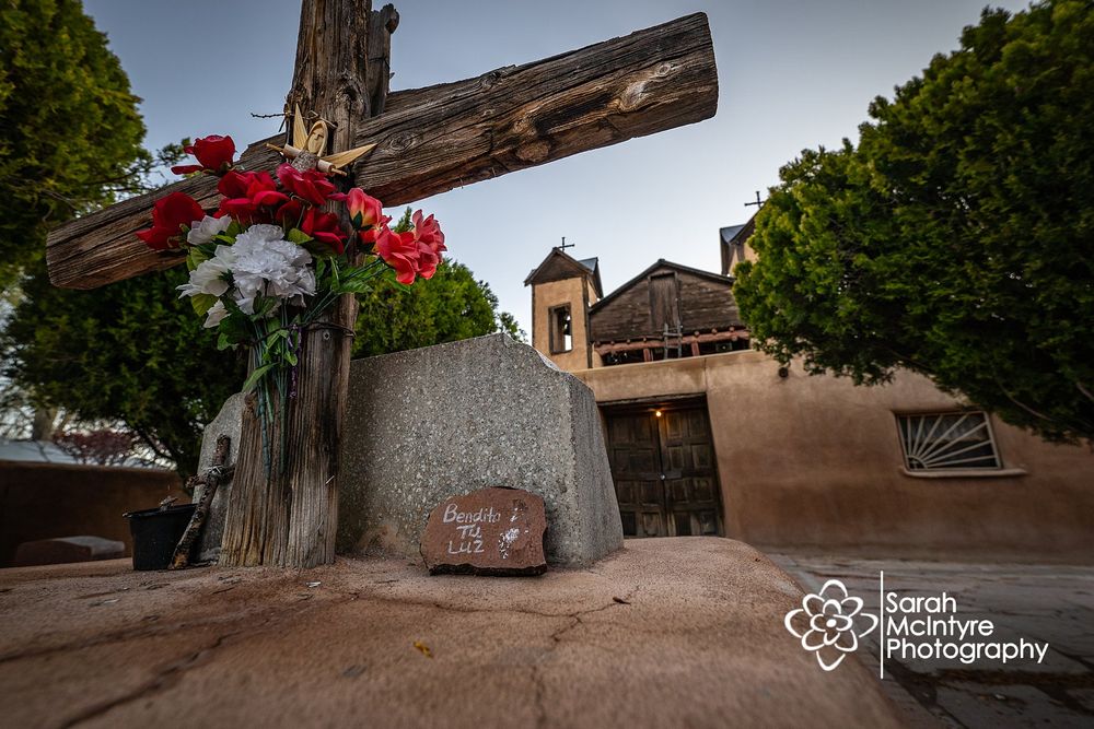 Wooden cross with red flowers in front of historic adobe church at sunset.