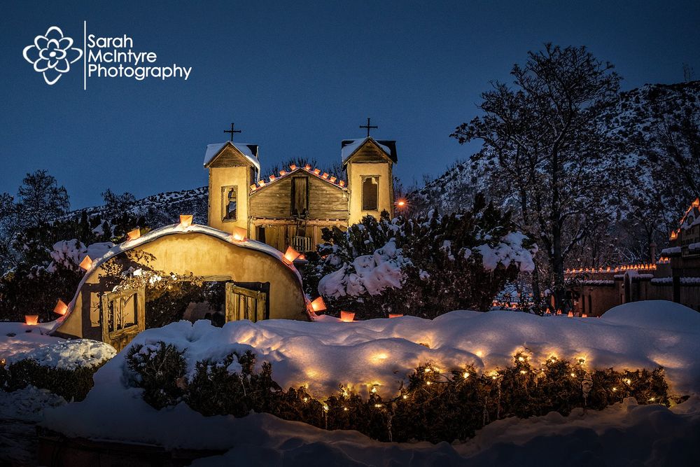 Historic adobe church illuminated with Christmas lights in snow at dusk against dark sky.