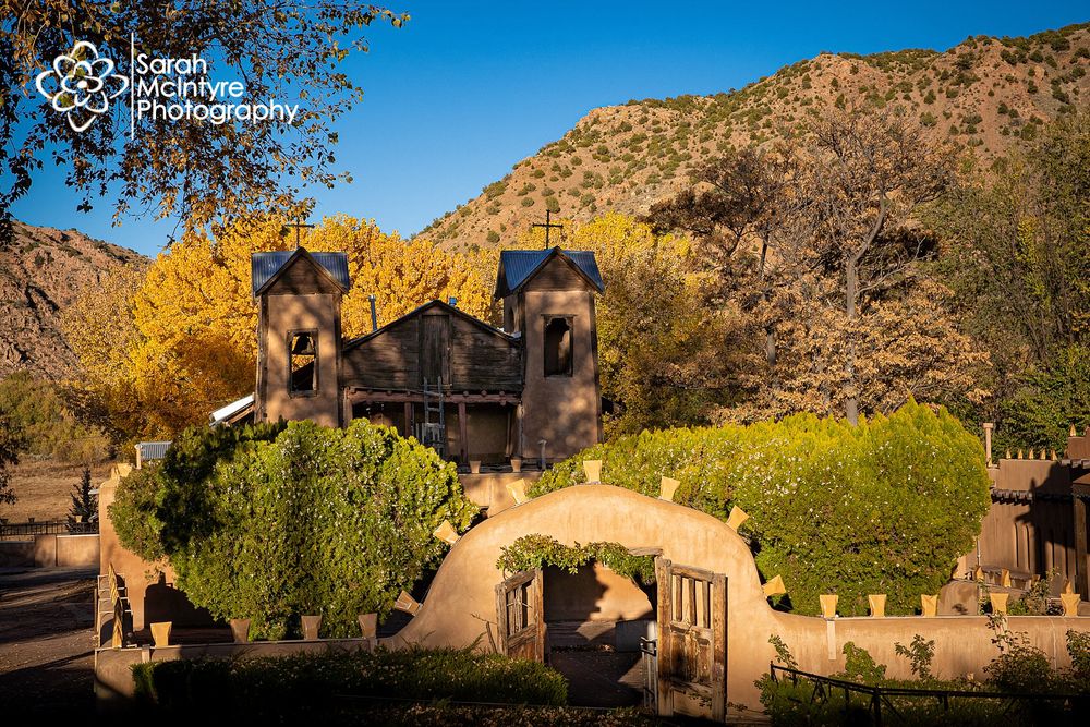 Adobe church with twin bell towers set against golden desert mountains and blue sky.