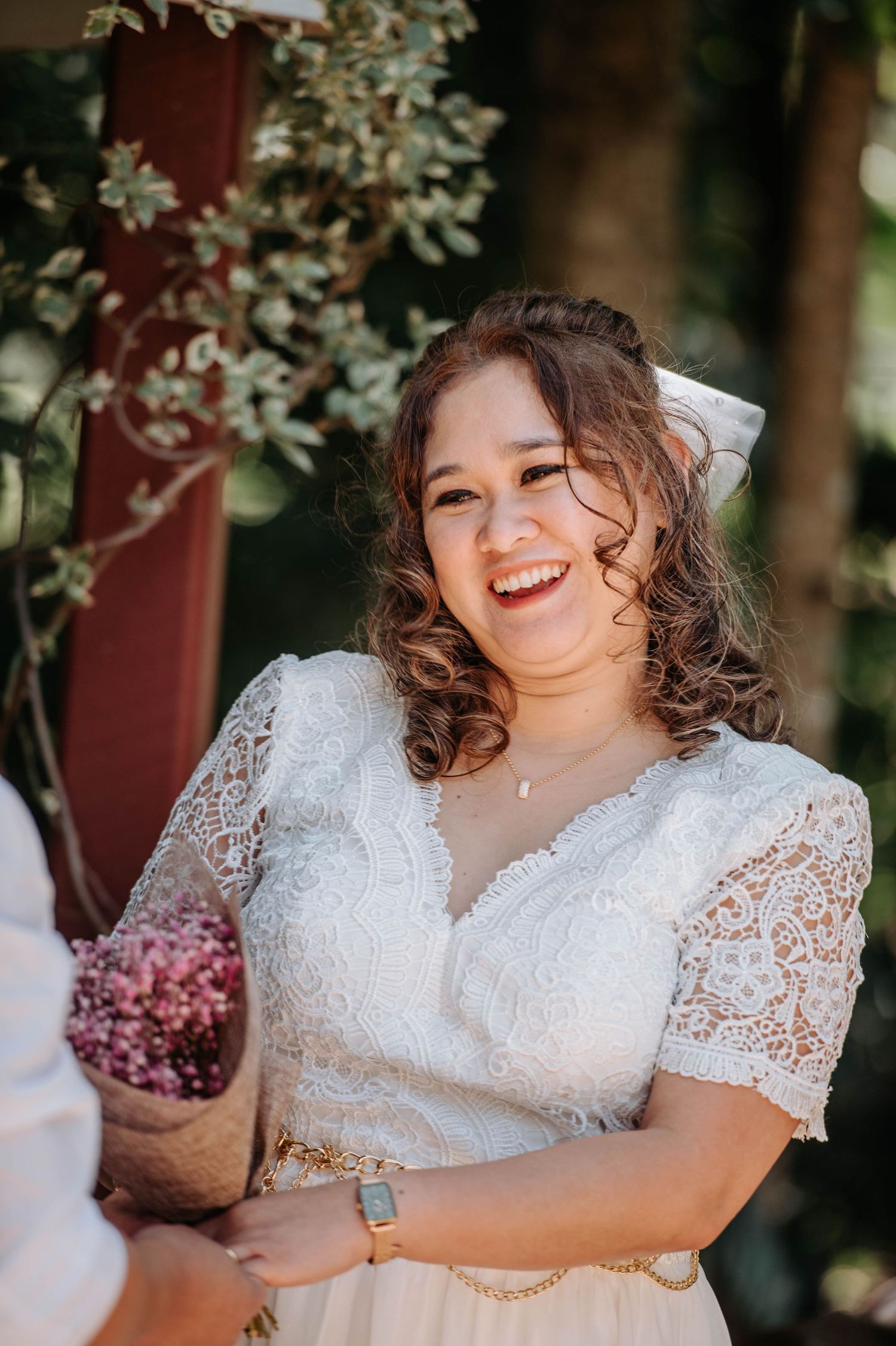 Bride in lace wedding dress smiles joyfully during outdoor ceremony.