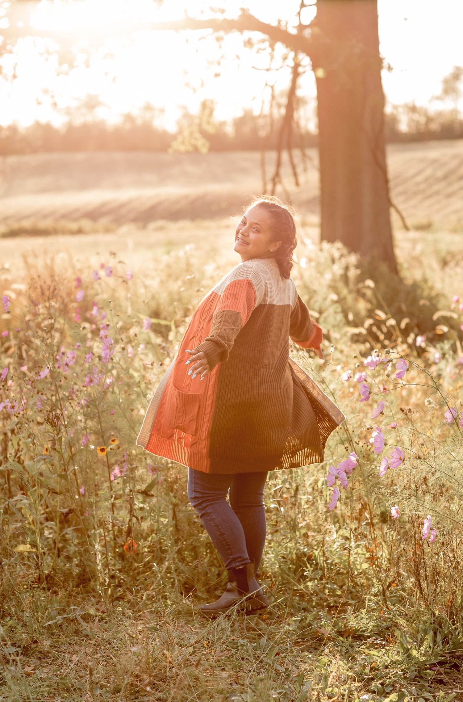 Young woman in colorful cardigan twirls joyfully in a sunlit wildflower meadow at sunset.