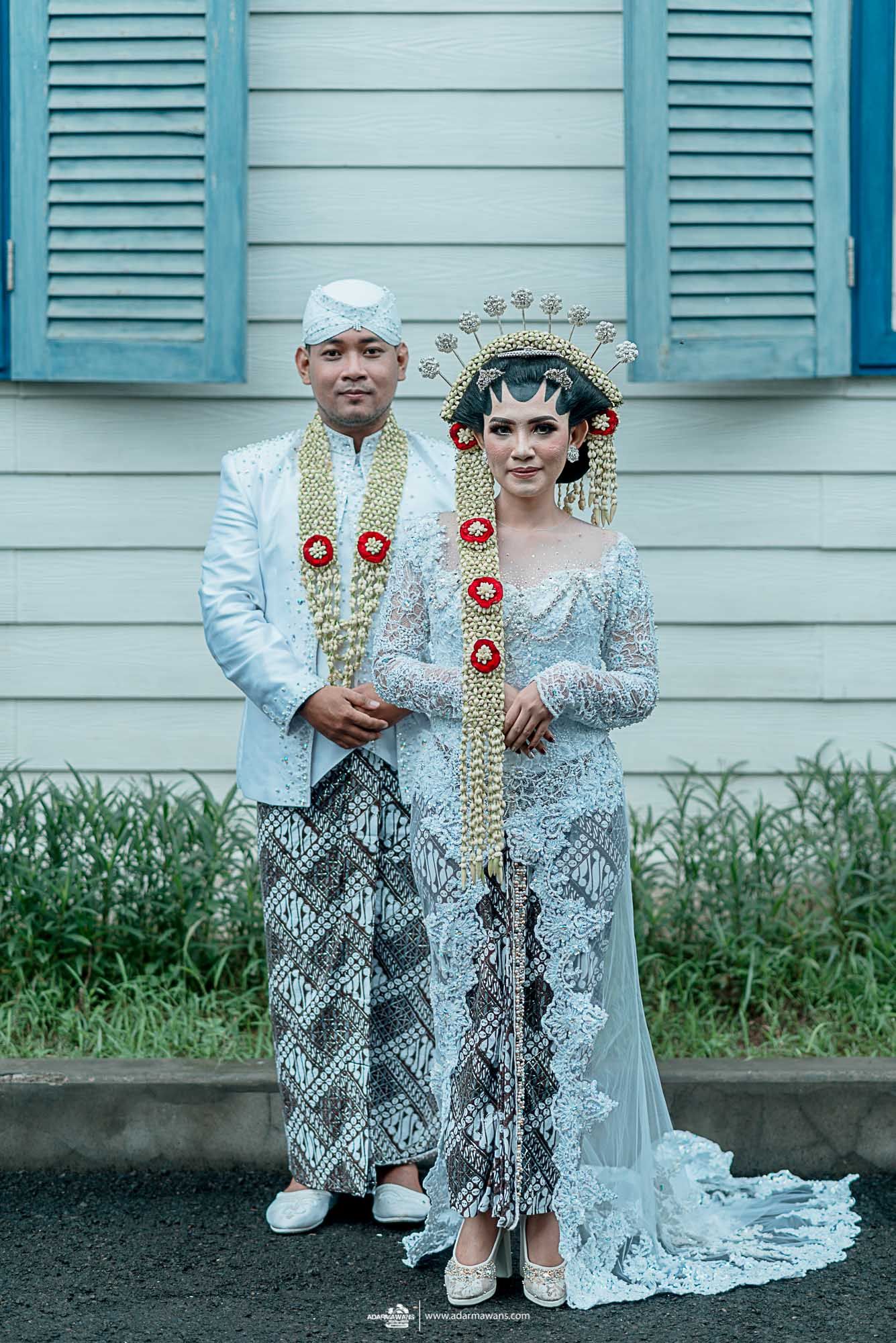 Traditional Indonesian wedding couple in light blue and white attire posing in front of a blue house.