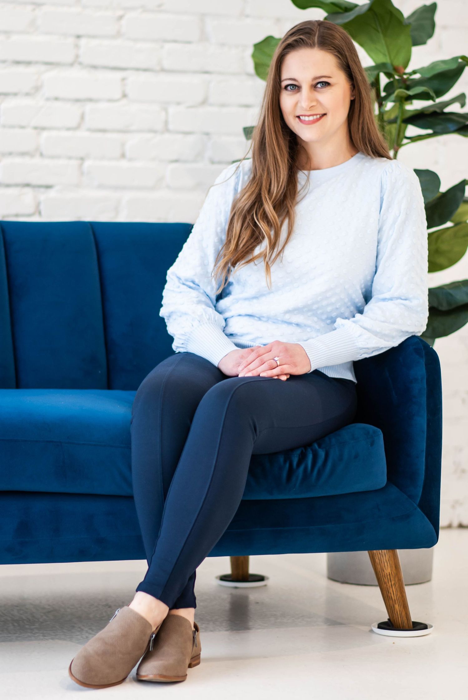 Woman in light blue sweater and navy leggings sitting on a blue velvet sofa smiling.