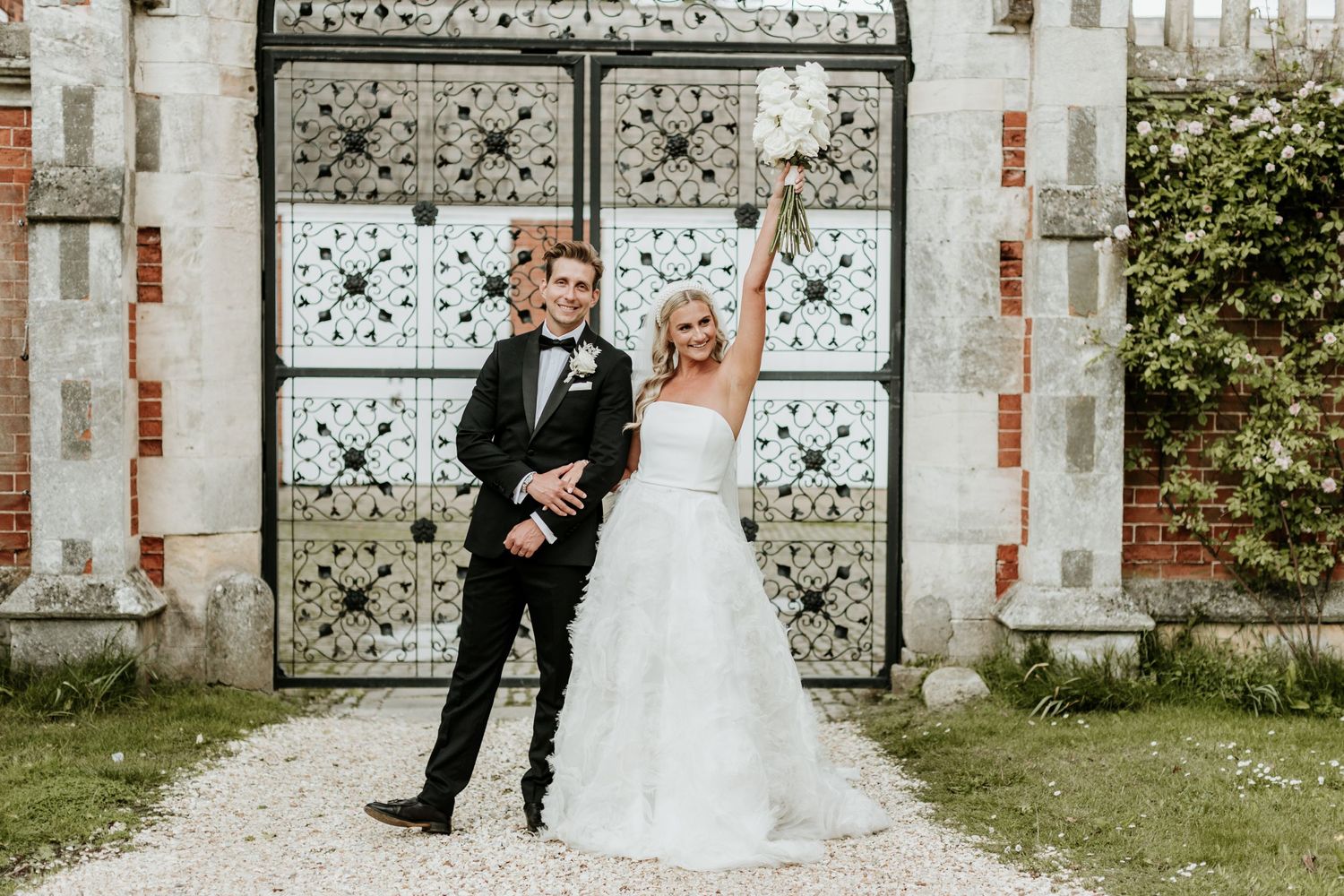 Wedding couple celebrates in front of ornate white gates.