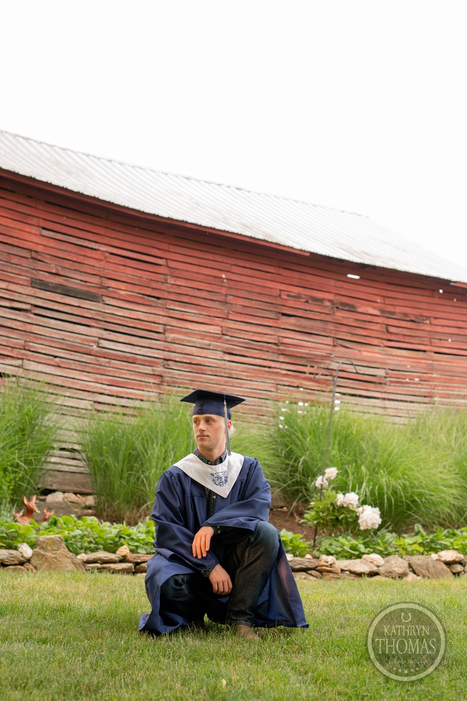 Graduate in academic regalia sits in front of a rustic red barn with ornamental grasses.