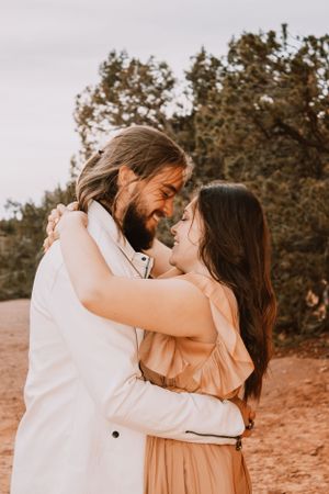 Couple sharing an intimate embrace in a desert setting with lush green trees in the background.