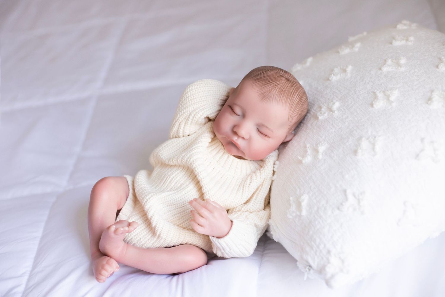 Baby doll laying on white blanket with pillow under head and cream sweater