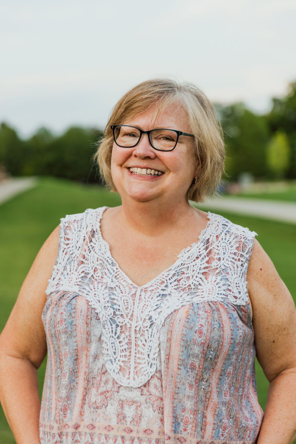 A person in a lace-trimmed top smiles warmly while standing in a green outdoor setting.
