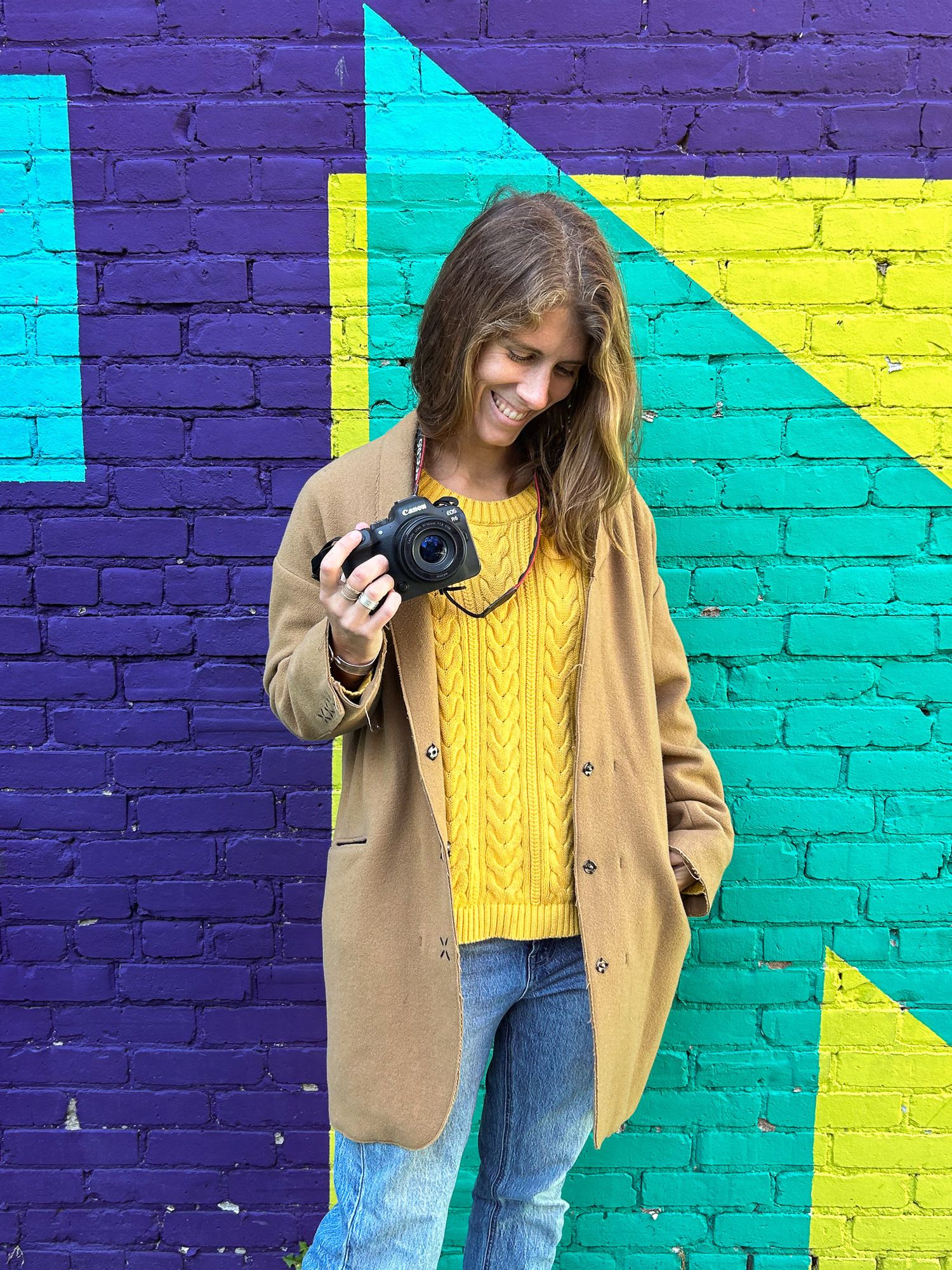 A photographer with long hair wearing a tan coat and yellow sweater poses against a colorful geometric wall mural.