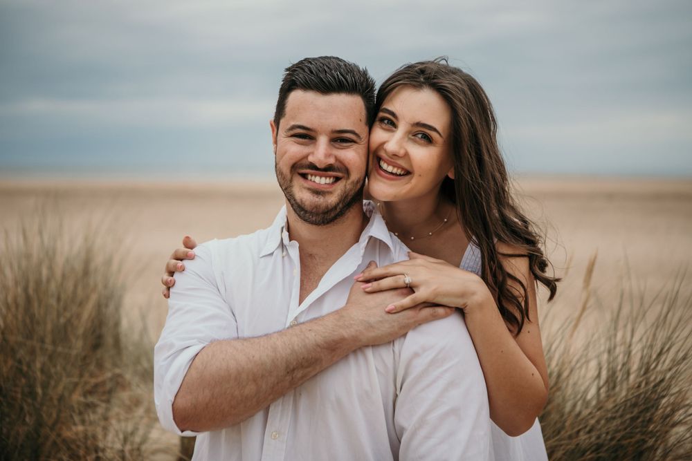 A couple in white outfits sharing an intimate moment on a beach with cloudy skies.
