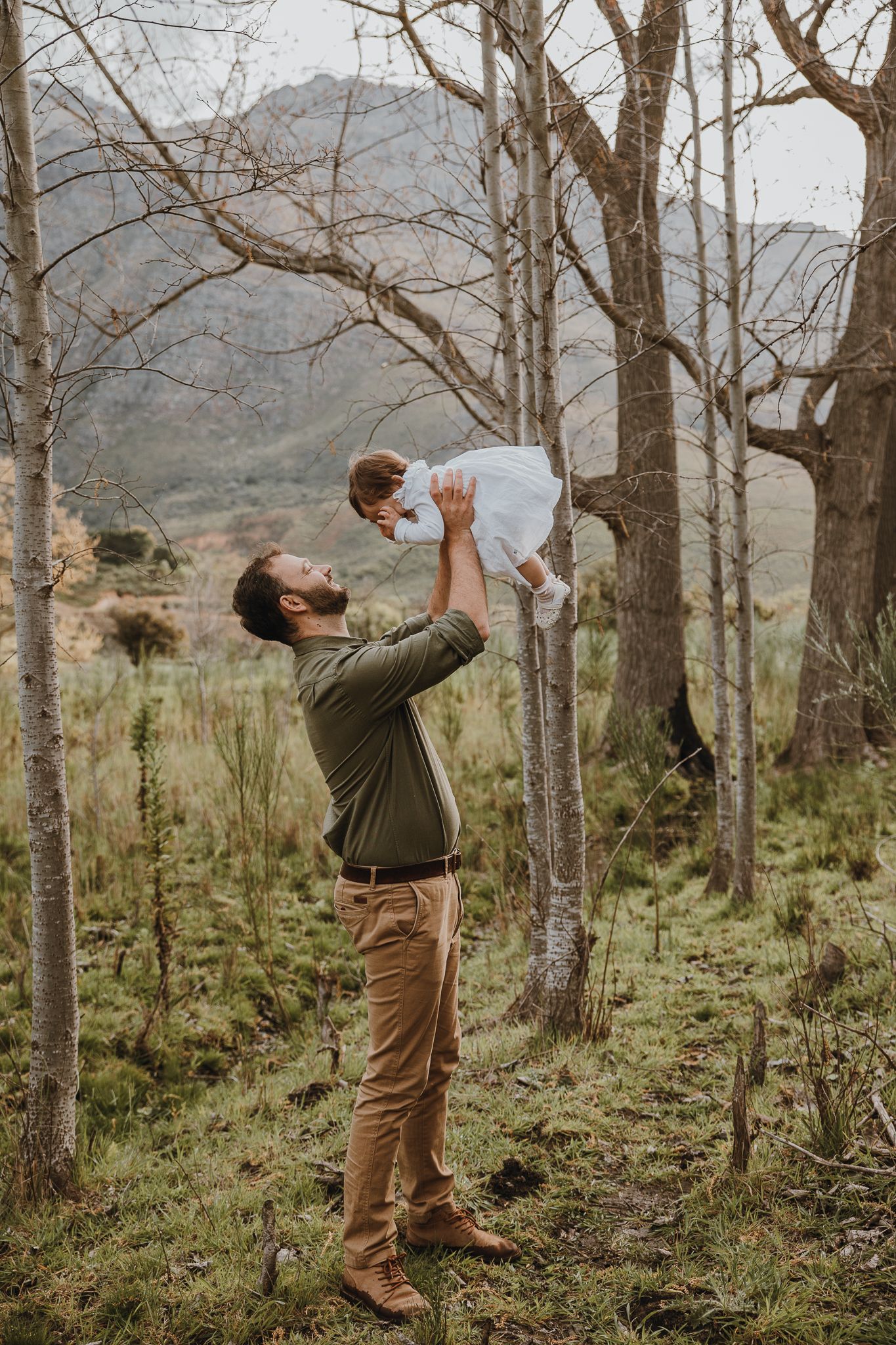Parent lifting child playfully in wooded mountain setting.