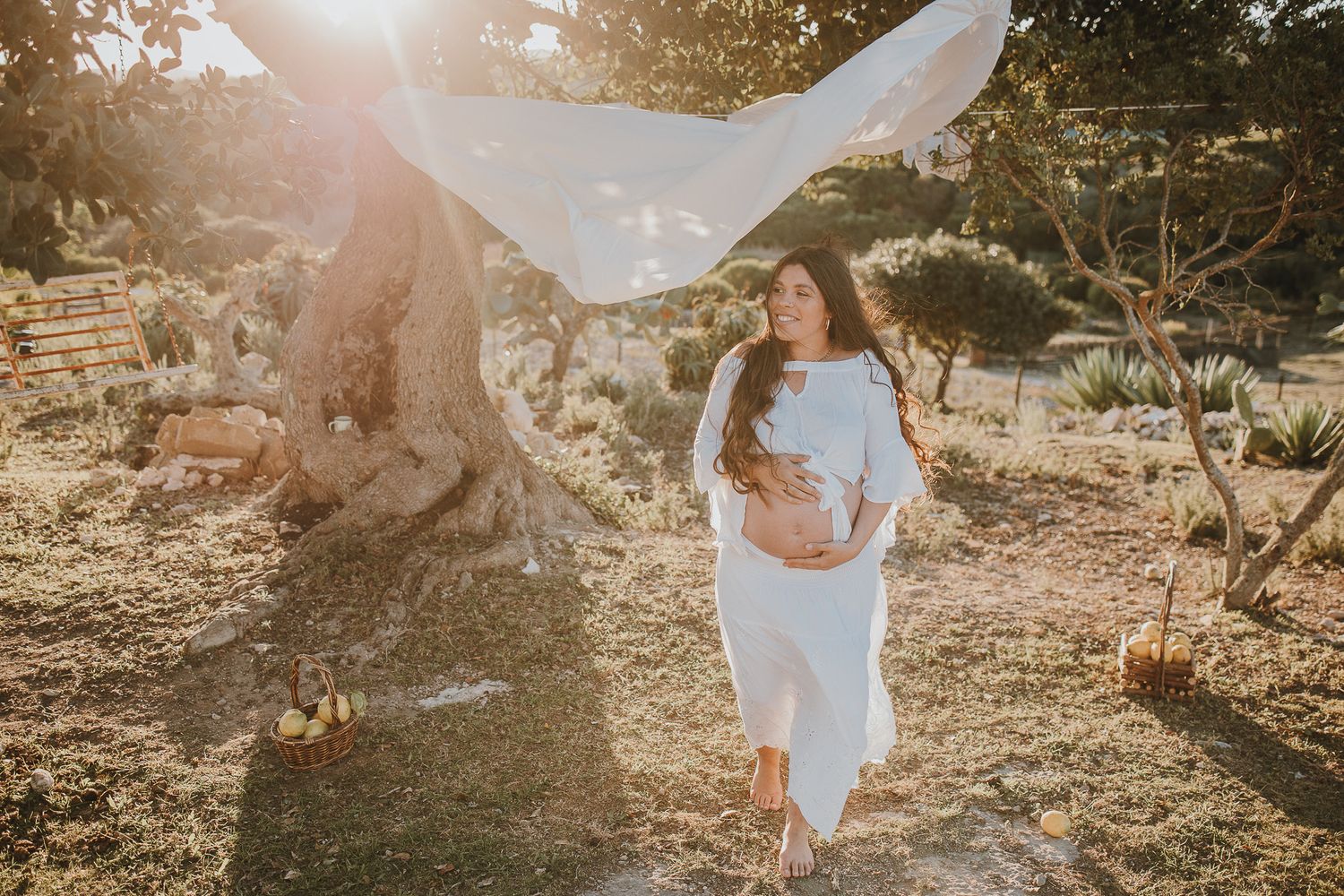 Maternity photo with flowing white fabric in golden sunlit outdoor setting.