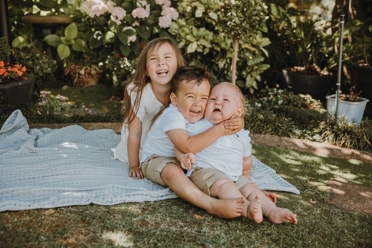 Three children share laughs while sitting on a blanket outdoors.