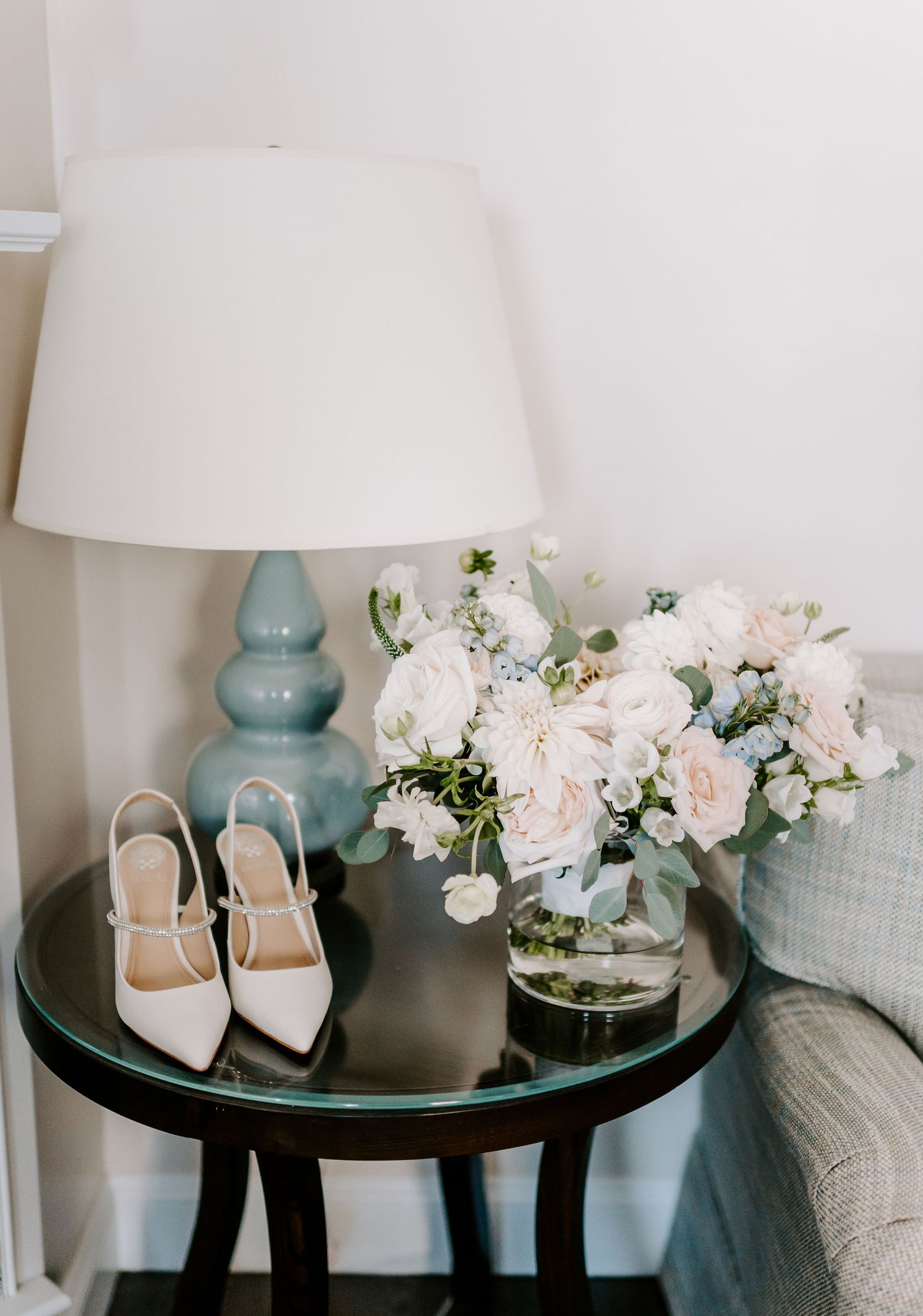 White bridal shoes and floral arrangement displayed on dark side table with blue lamp.