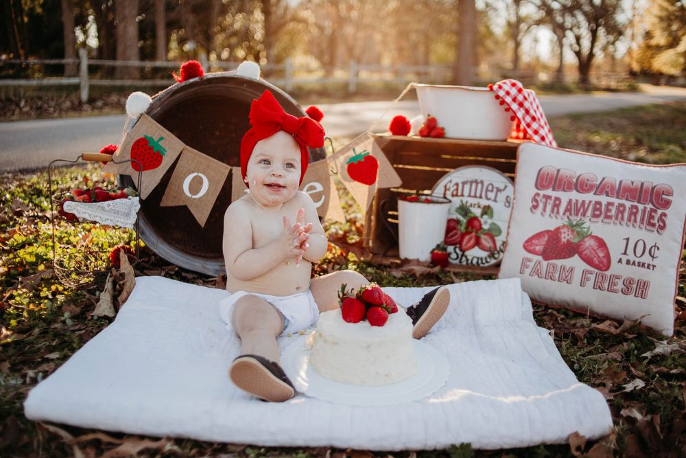 A Valentine's Day themed baby photo setup with strawberries, hearts, and rustic props on a white blanket.