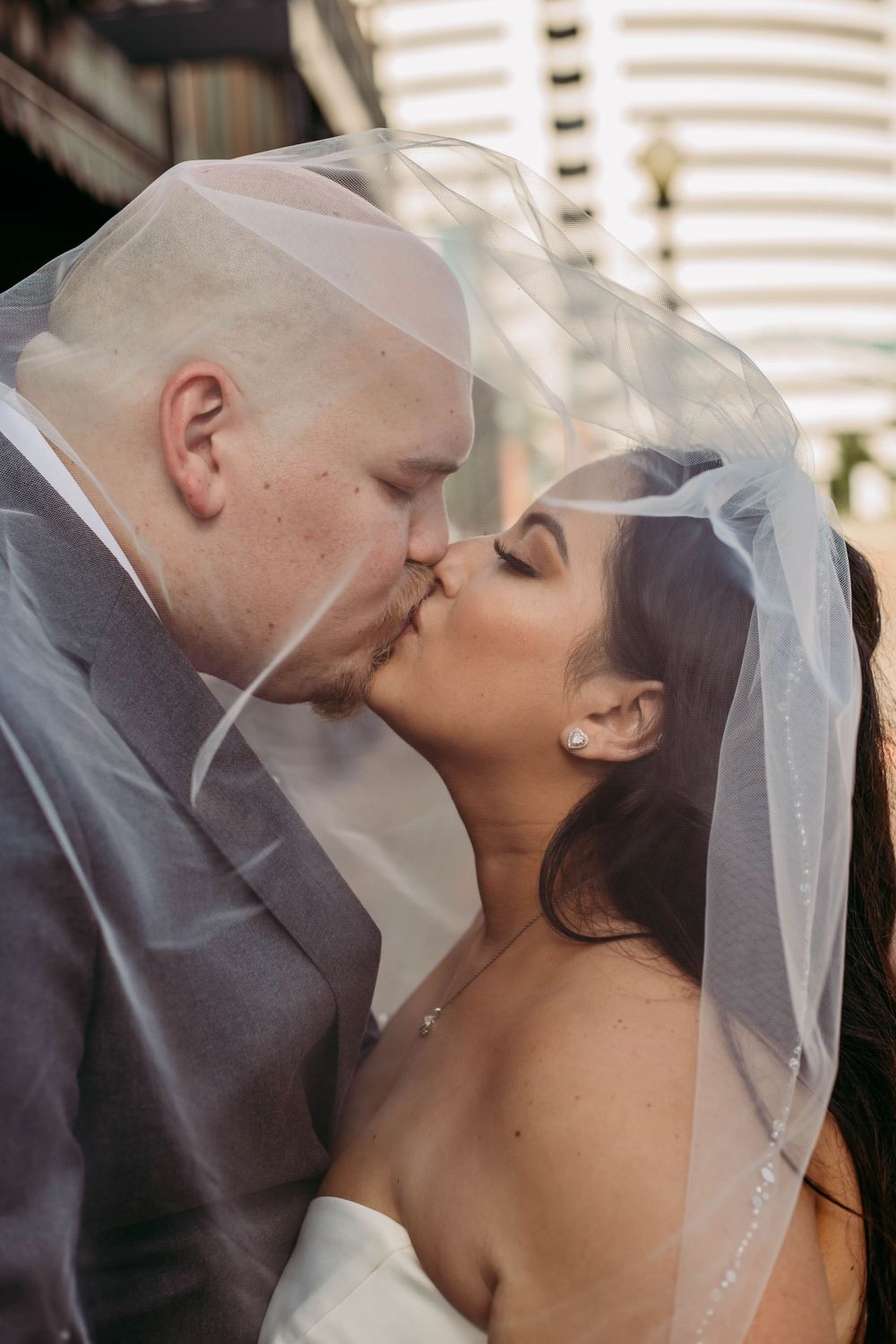 A tender wedding kiss between newlyweds under a delicate white veil against an urban backdrop.