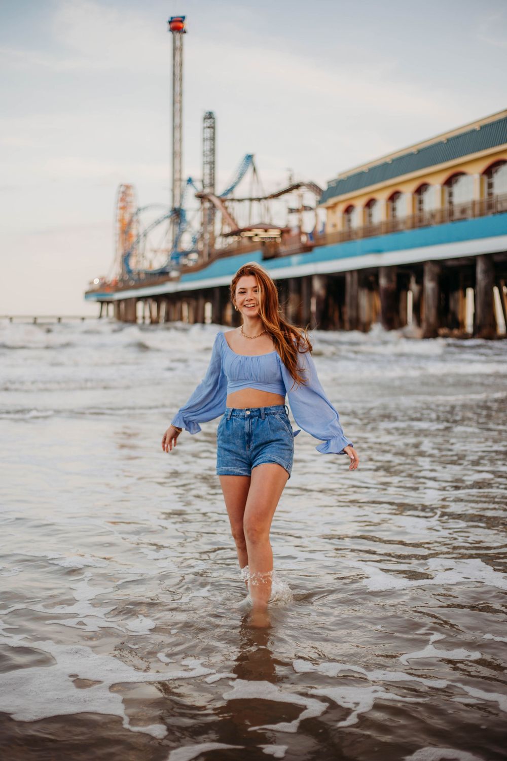 A woman walks through ocean waves near a pier with an amusement park in the background.