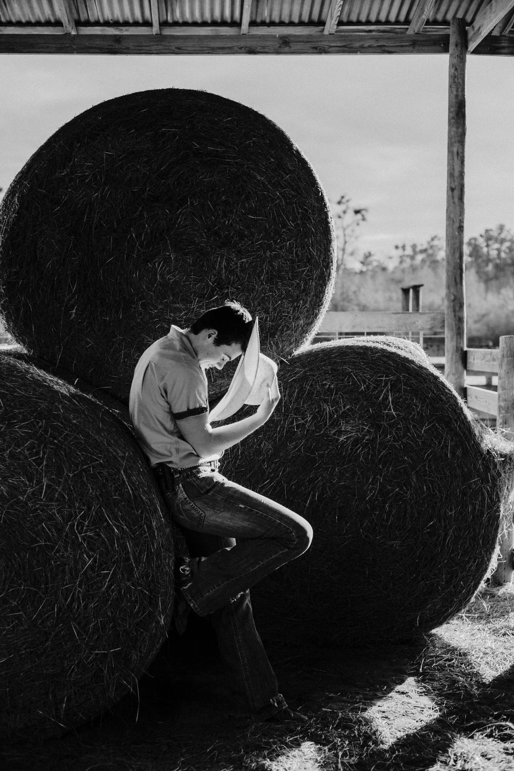 A black and white portrait of someone leaning against large round hay bales under a barn roof.