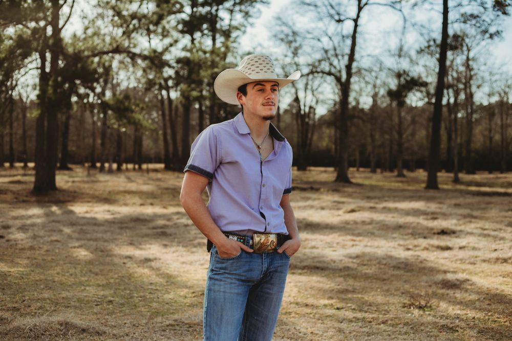 A casual portrait of someone in a cowboy hat and purple shirt against a rustic wooded background.