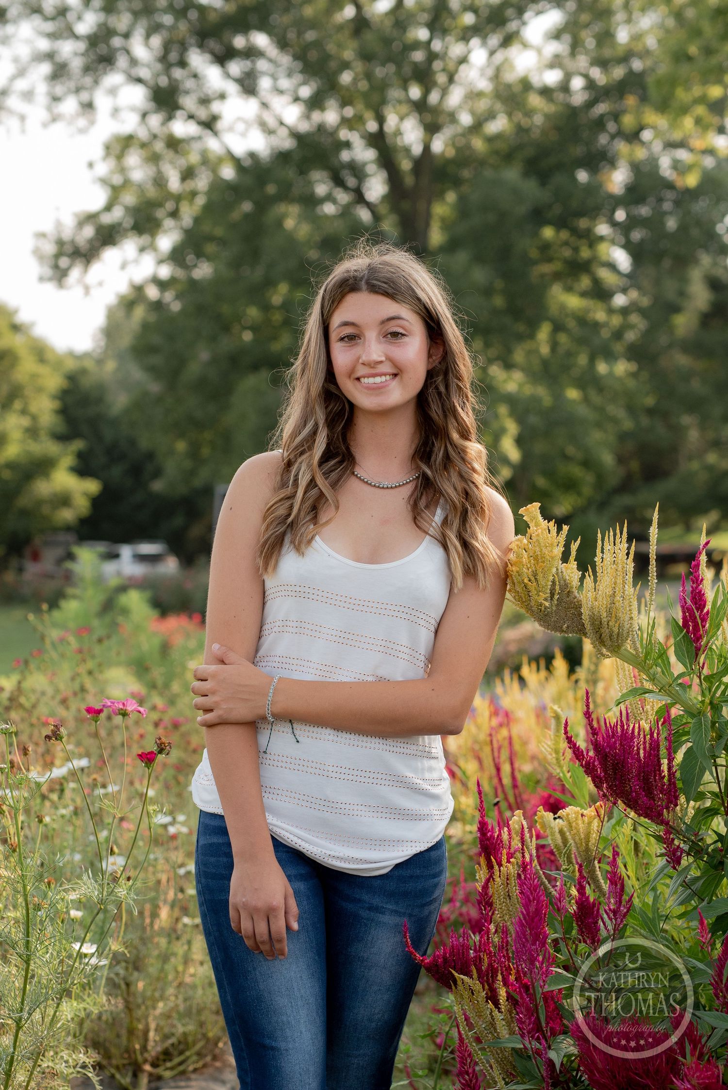 A person in a white tank top stands among colorful summer flowers with a farmhouse in the background.