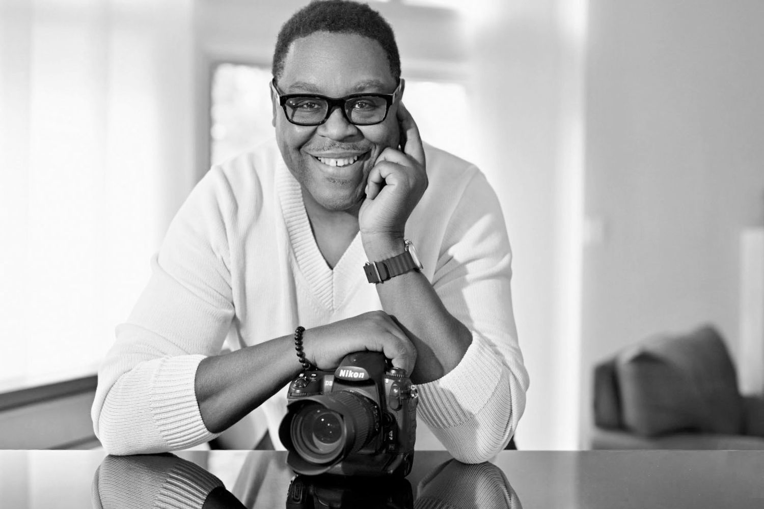 Black and white portrait of smiling man with glasses leaning on table with Nikon camera.