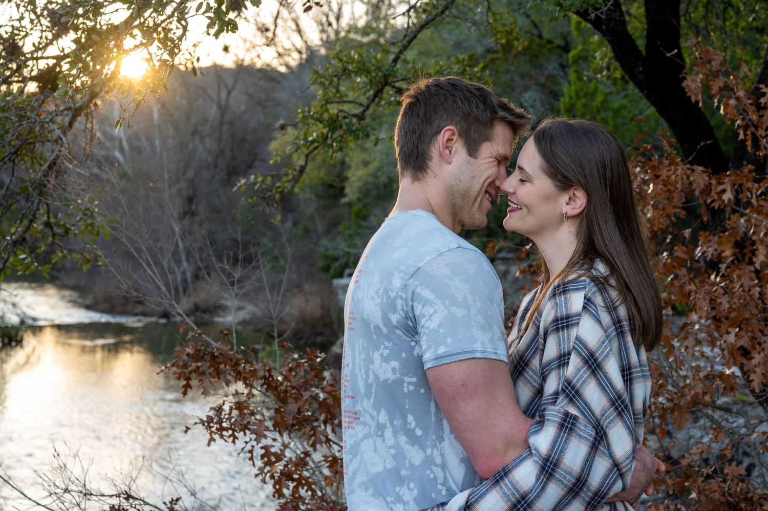 A romantic couple shares an intimate moment by a lake during sunset with fall foliage in the background.