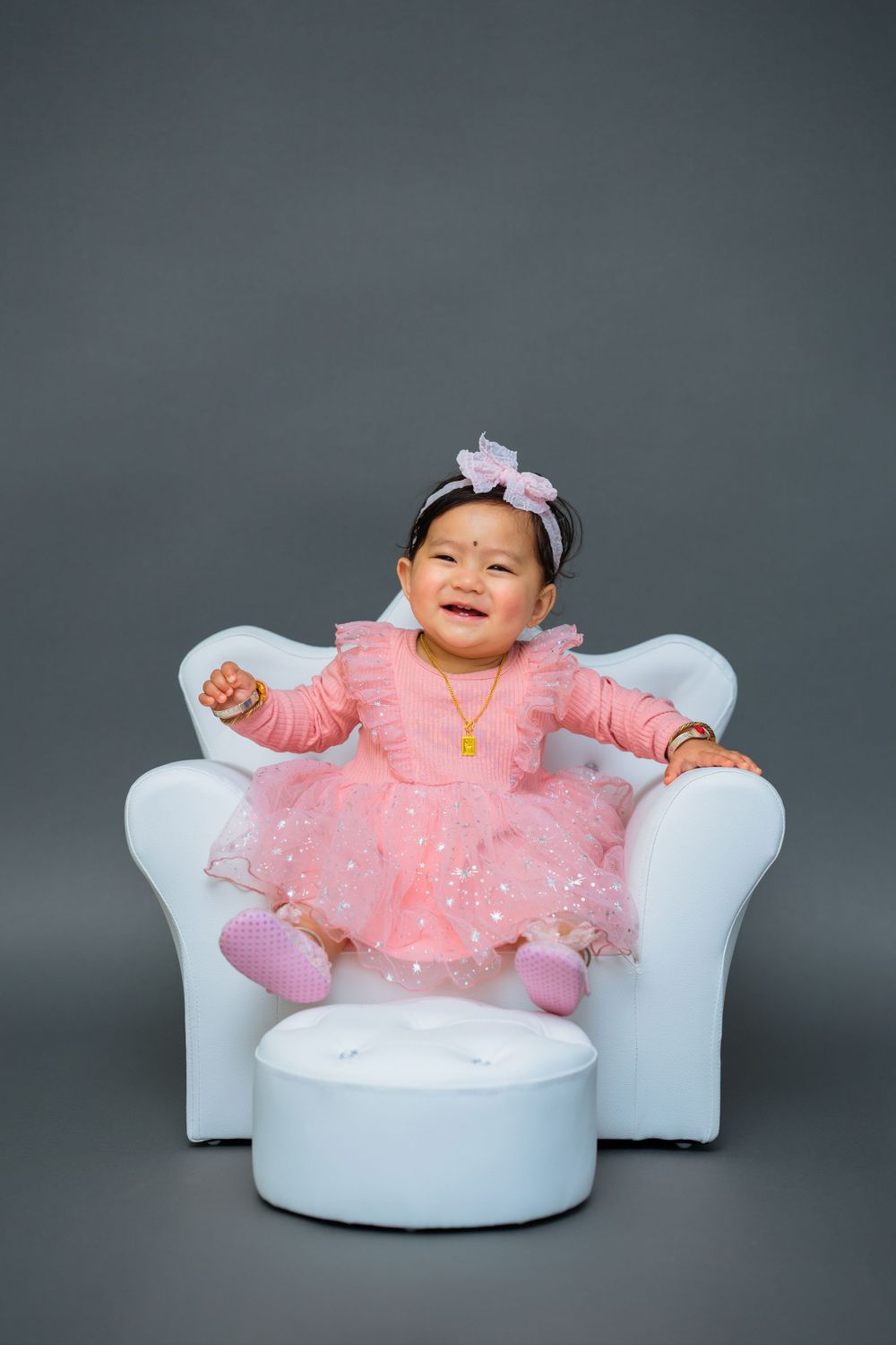 Smiling baby wearing pink tulle dress poses in white armchair against dark backdrop.
