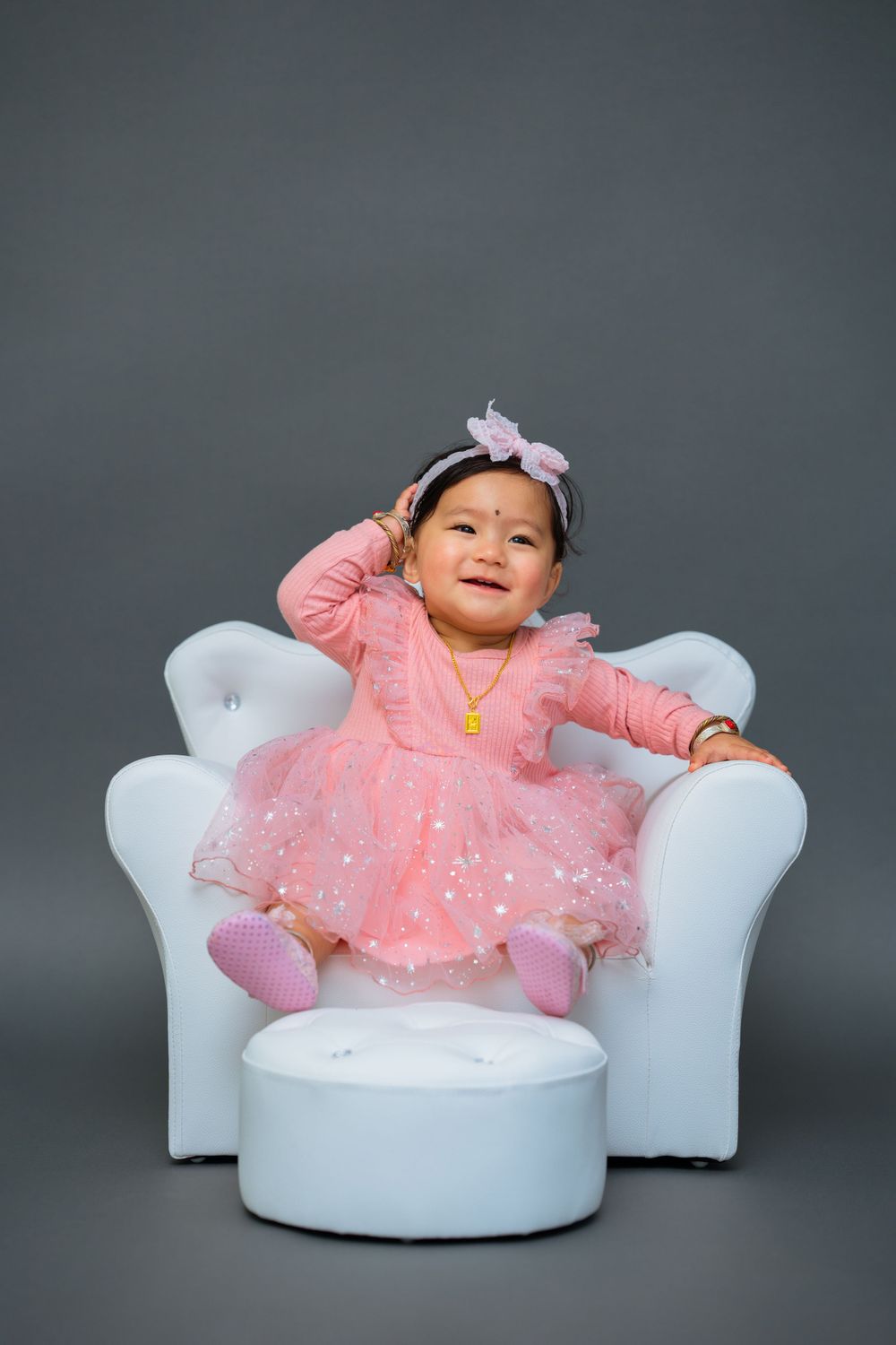 A small child wearing a sparkly pink dress and bow sits cheerfully on a white tufted chair with ottoman against grey backdrop.