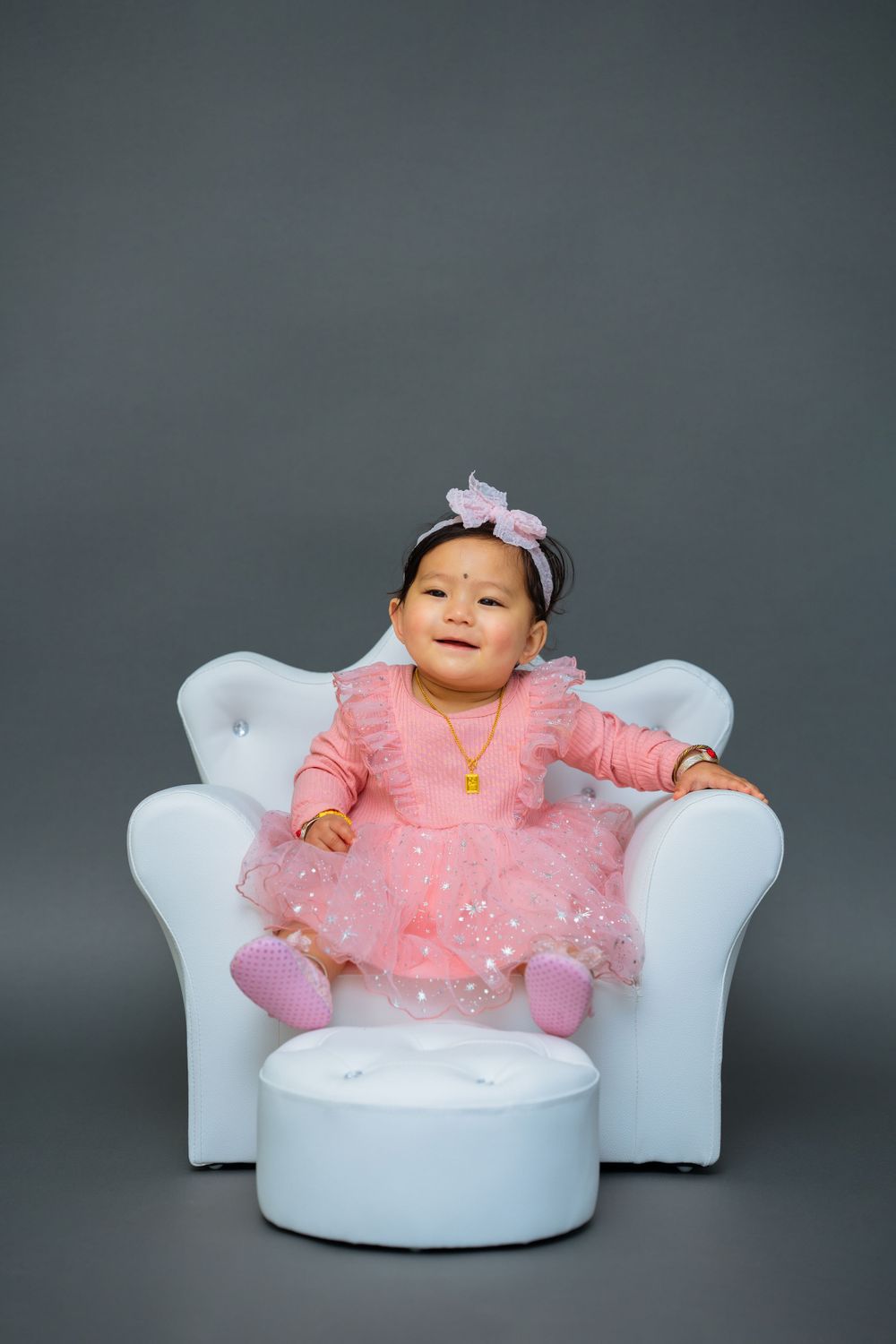 A toddler in pink tulle dress and headband relaxes on an elegant white chair with matching footstool.