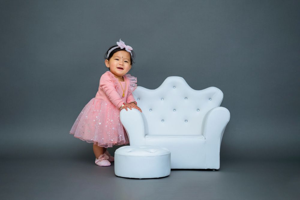 A baby in a pink party dress stands beside a white tufted chair and ottoman in a studio setting.