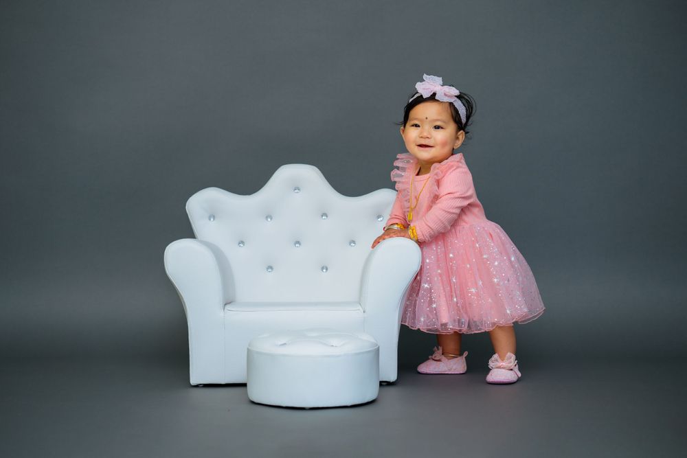 A little one in a pink tulle dress poses next to a white tufted chair in a professional photo studio.
