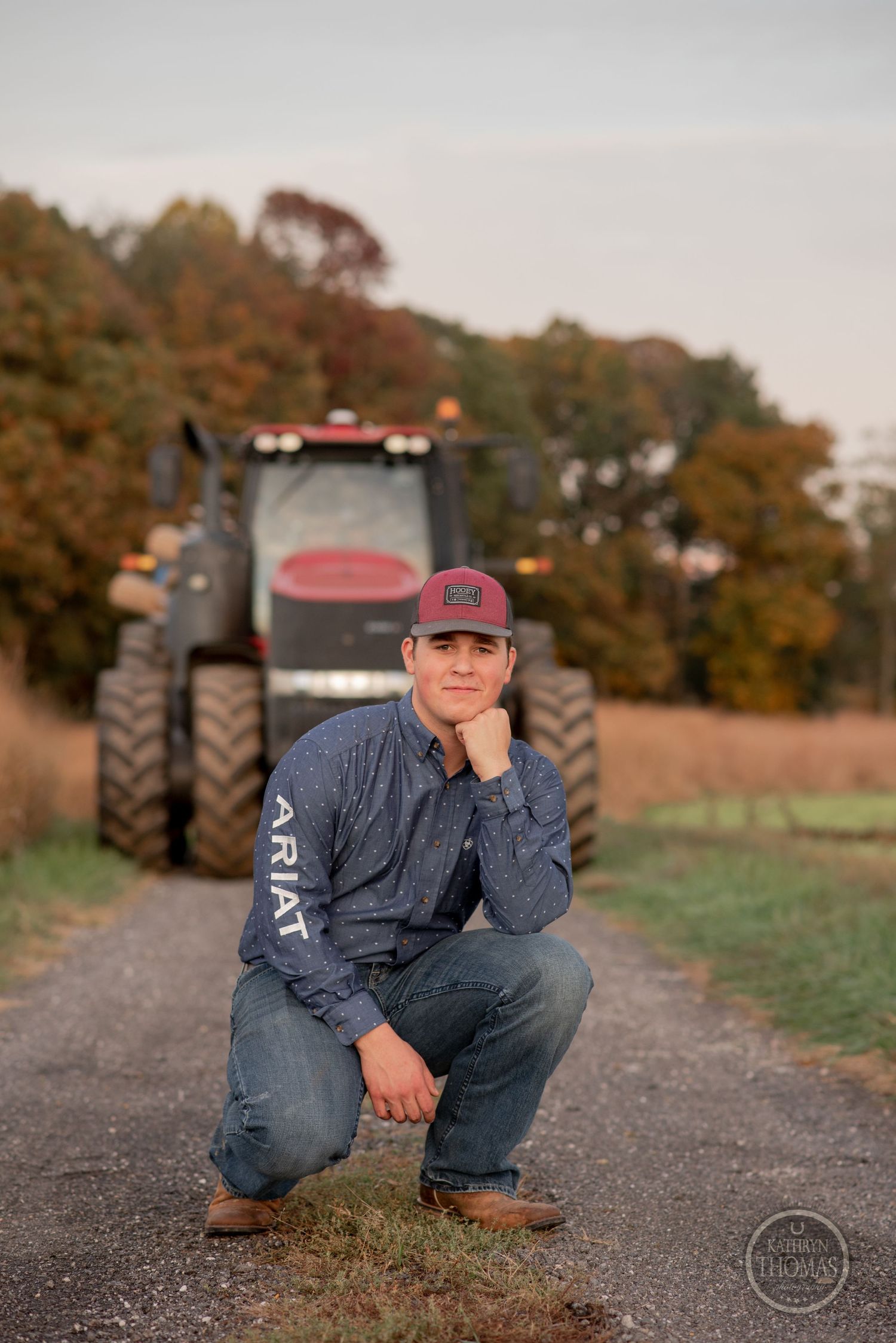 Person crouches on dirt path with large farm tractor and fall trees in background.